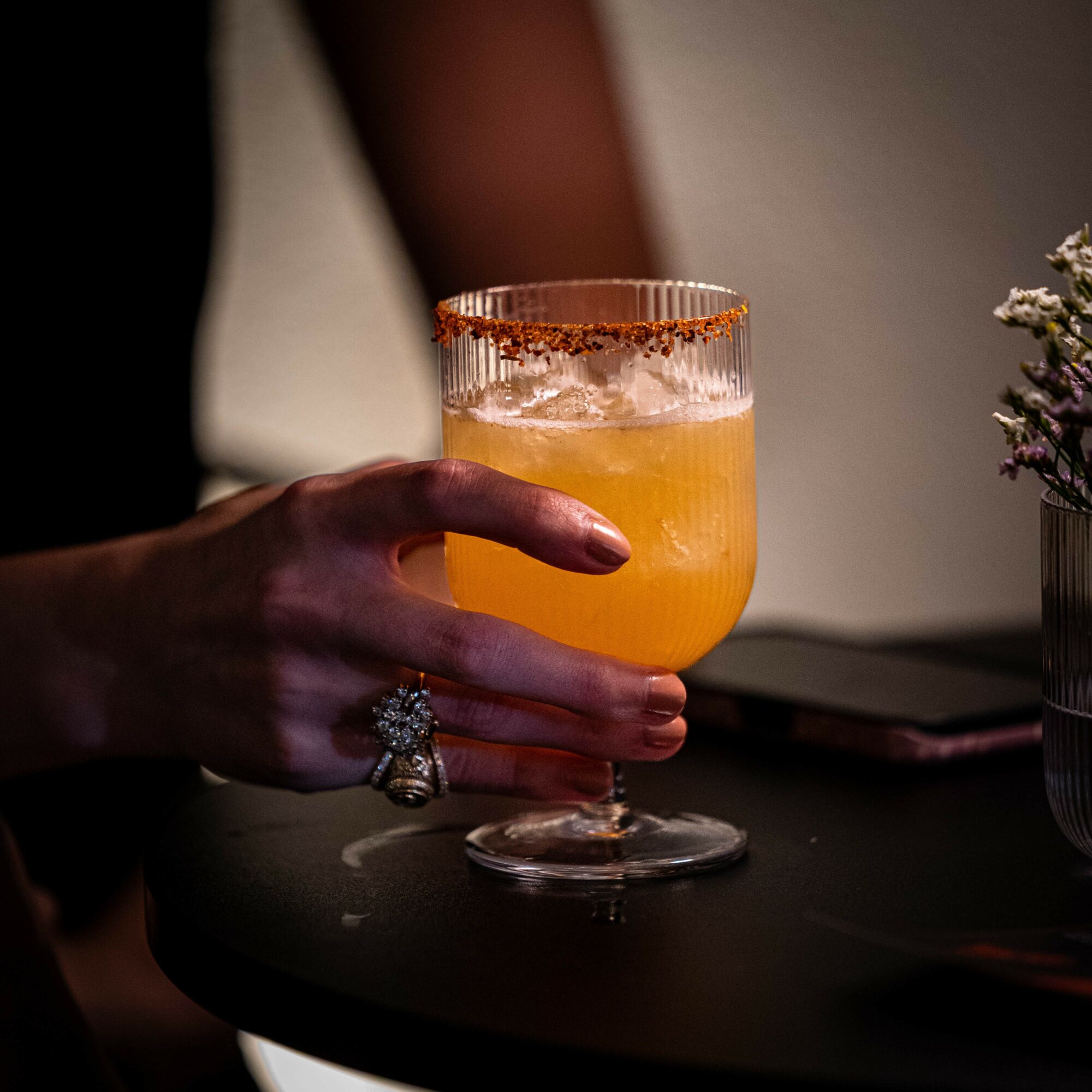 Person holding a glass with orange-colored drink, decorated with a salted rim, on a dark table.
