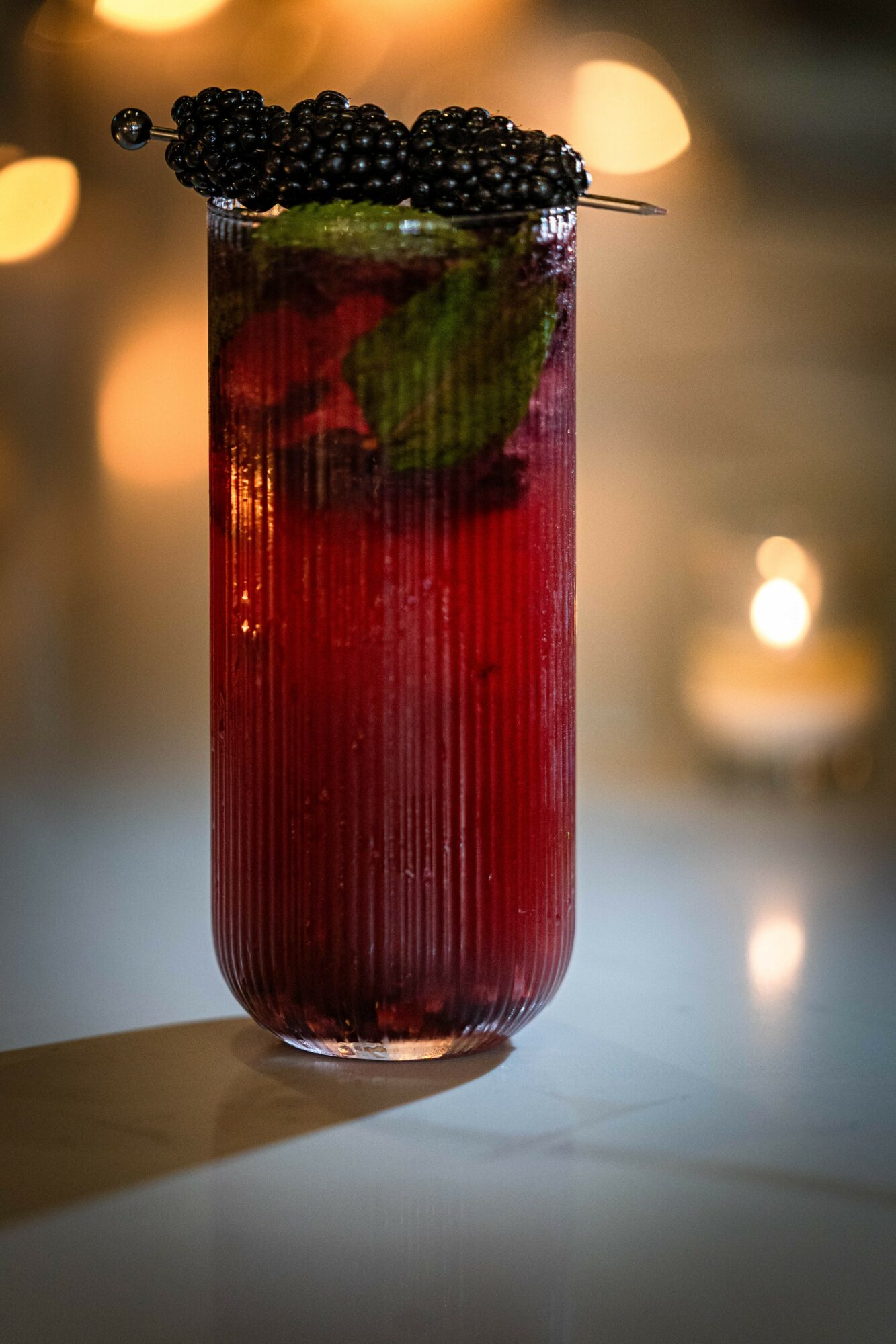 Tall glass with dark red drink, garnished with blackberries and mint, on a table with blurred candlelight background.
