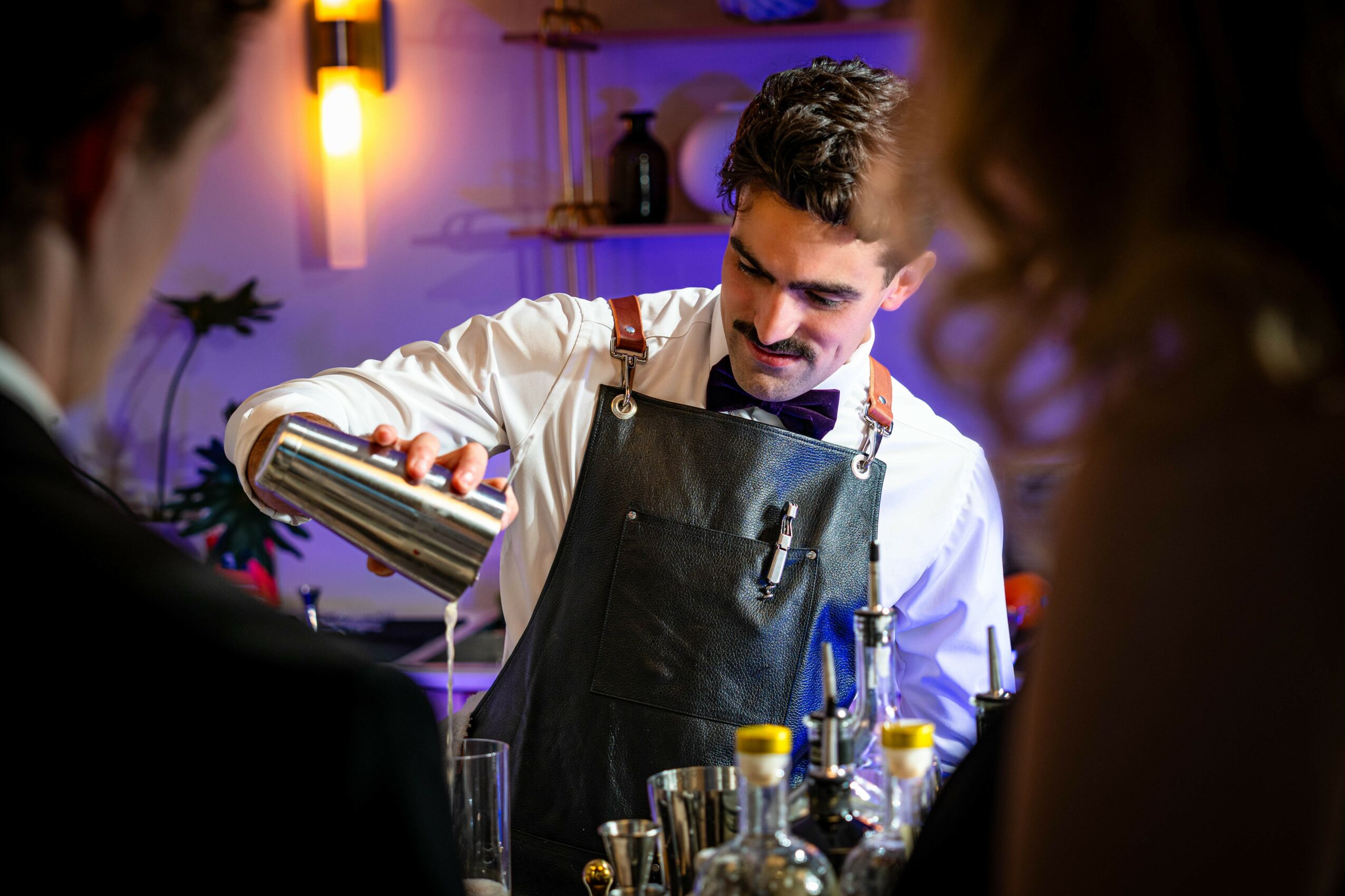 Barista pouring a drink in a cafe with glasses and bottles on the counter, warm lighting in the background.