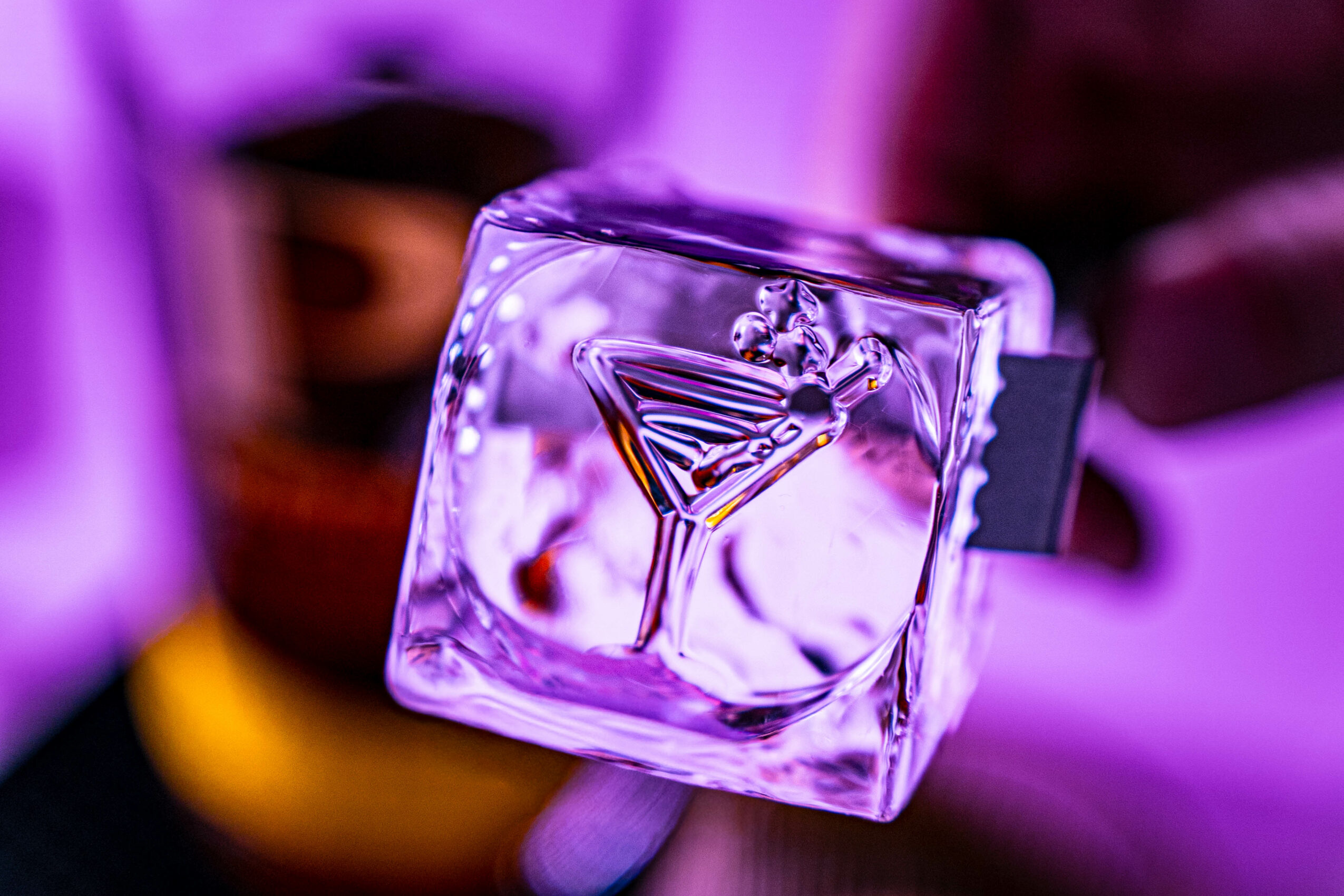 A glass with a martini inside, held by a hand, with a purple background and colorful lighting.