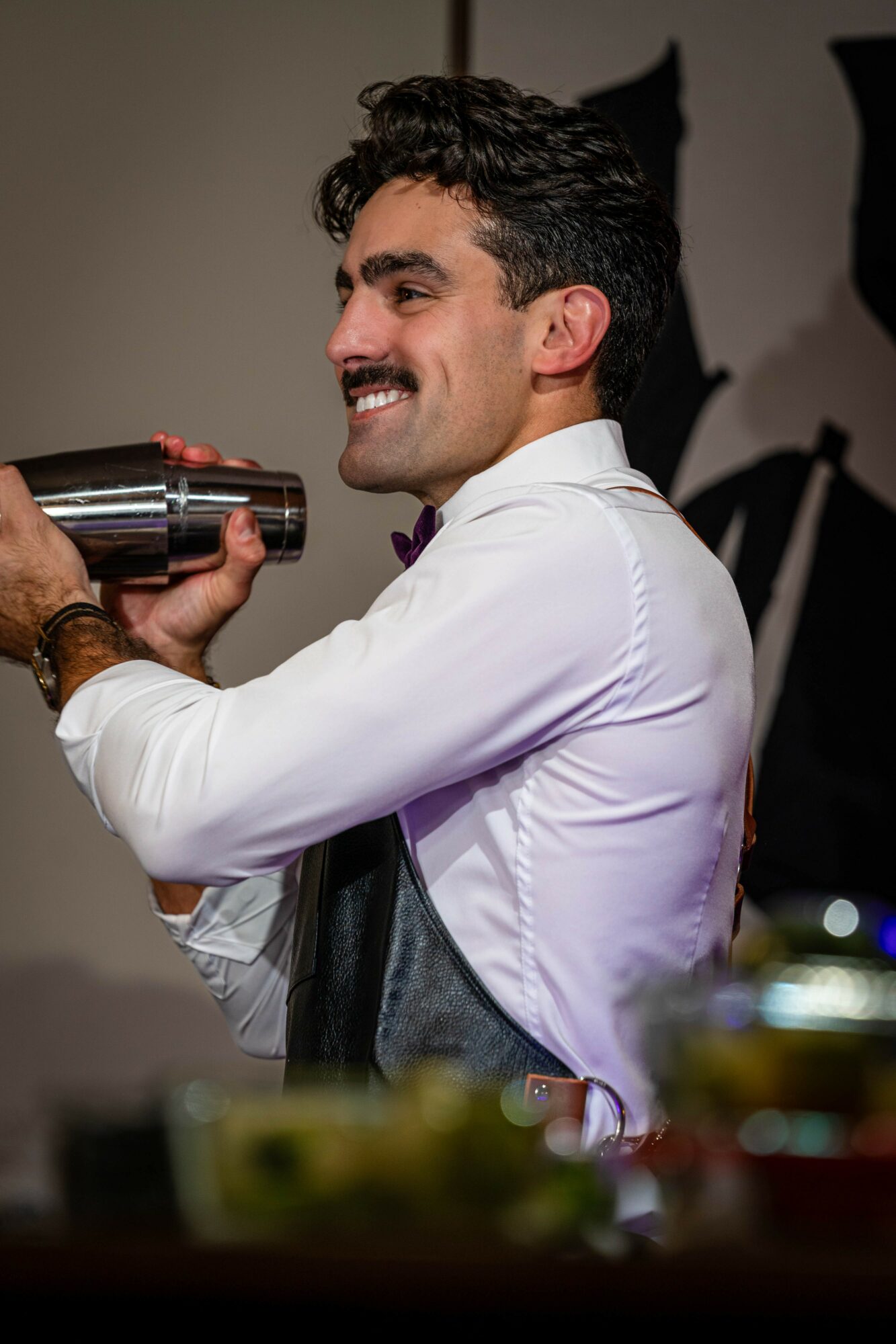 Man with dark curly hair and mustache smiling, holding a cocktail shaker, wearing a white shirt.