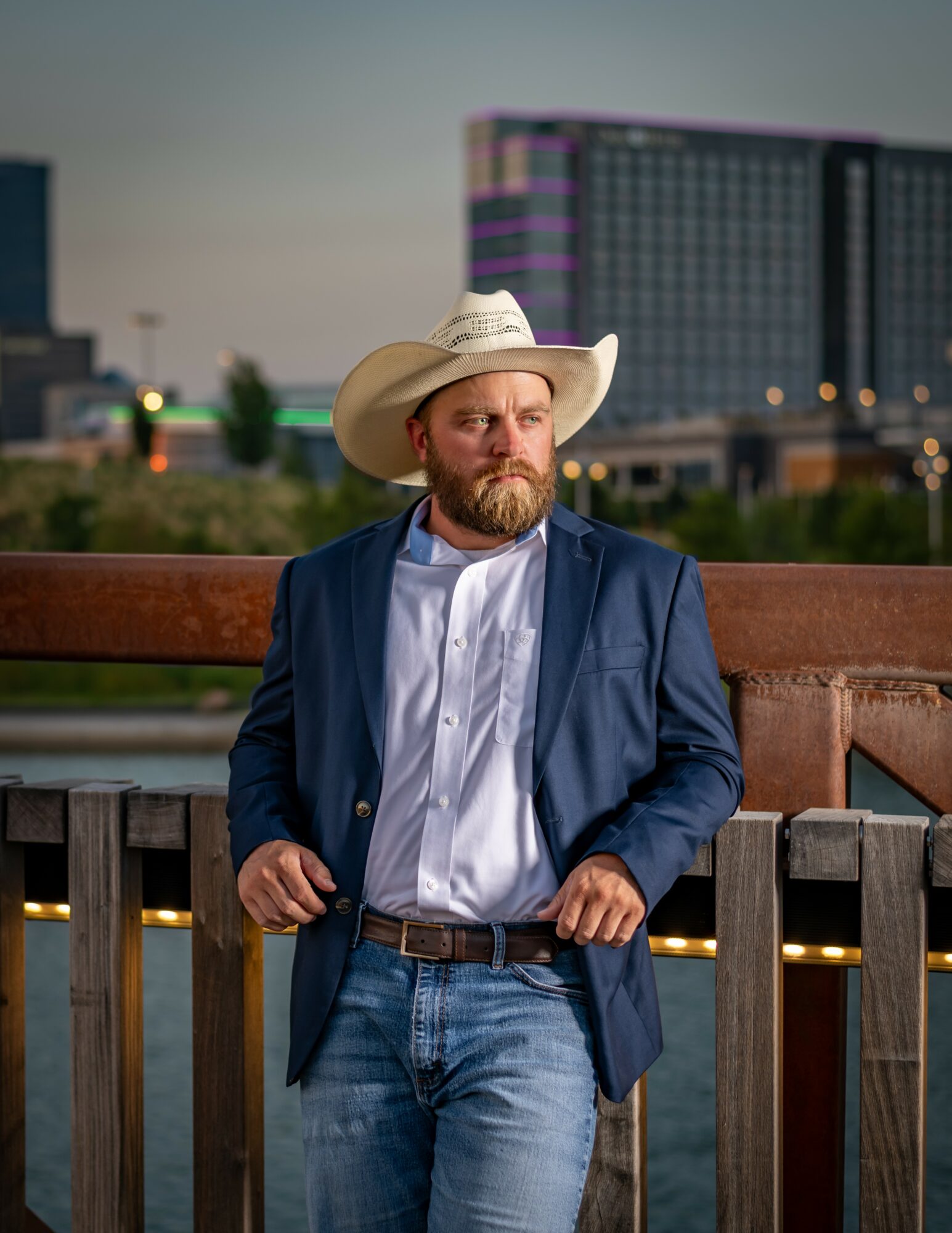 Man with a beard wearing a cowboy hat, blue blazer, white shirt, and jeans stands outdoors near a wooden railing with city buildings in background.