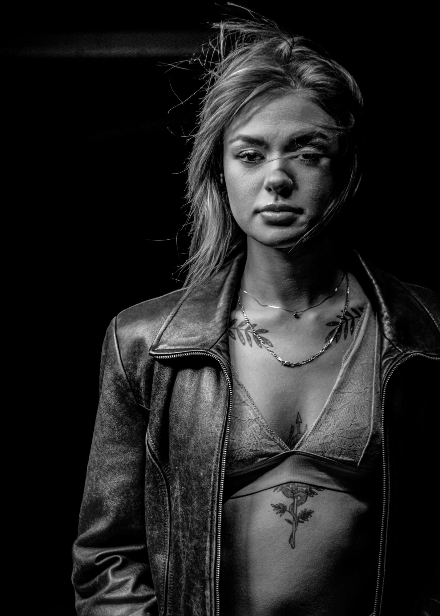 Young woman with tousled hair, wearing a leather jacket and a top revealing a tattoo on her chest.