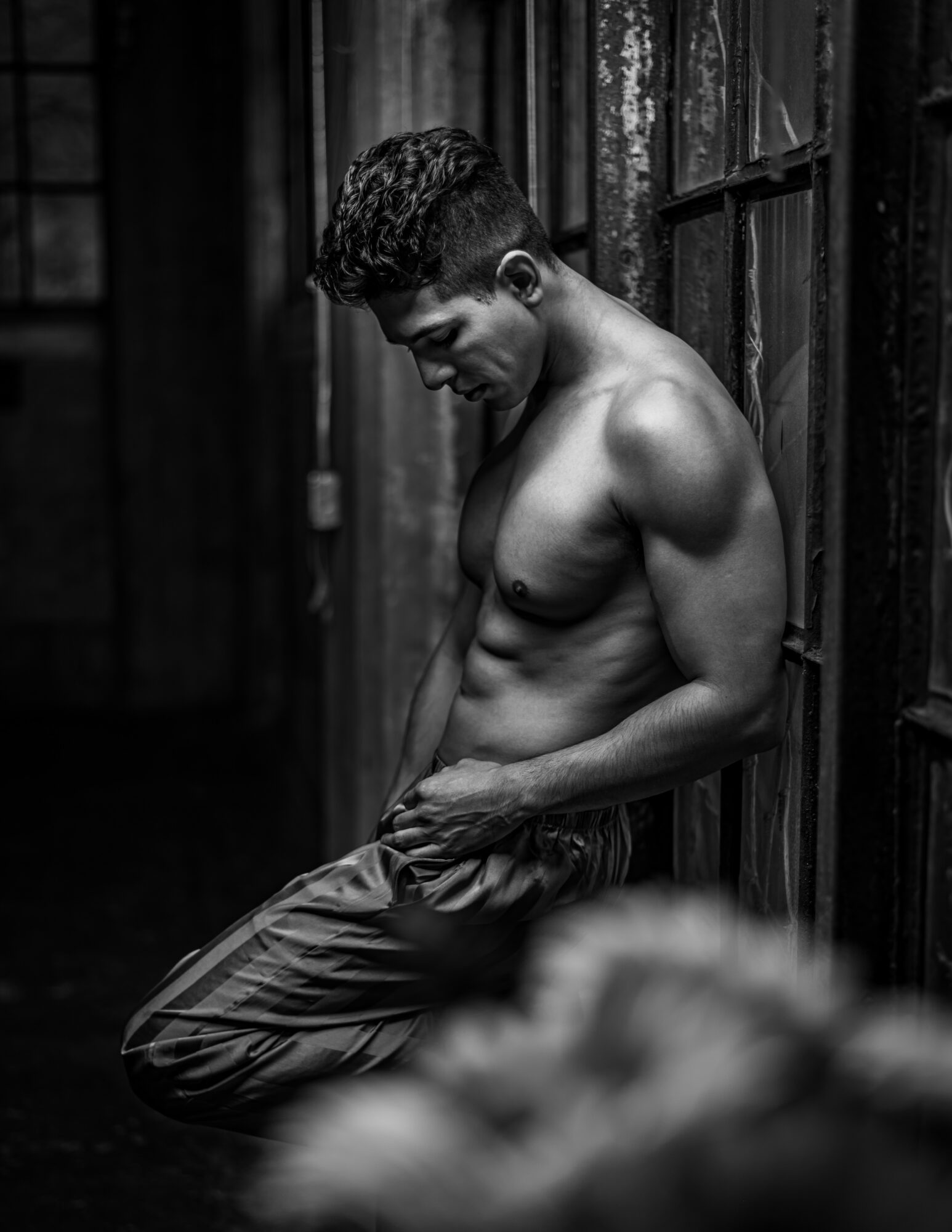 Young man with muscular build leaning against wooden wall, shirtless, in black and white photo.
