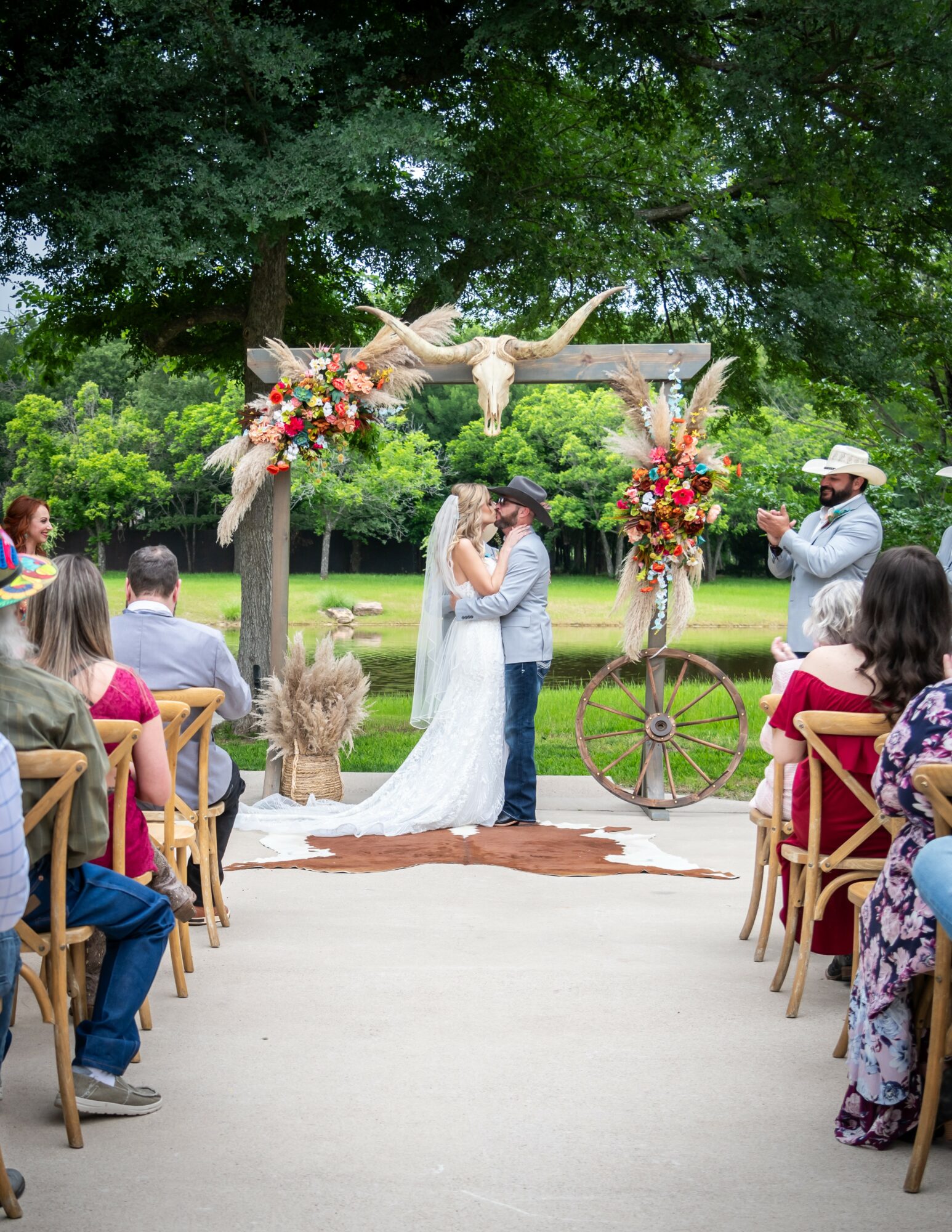 Bride and groom kissing under a decorated arch with flowers, guests seated outdoors, trees in background.