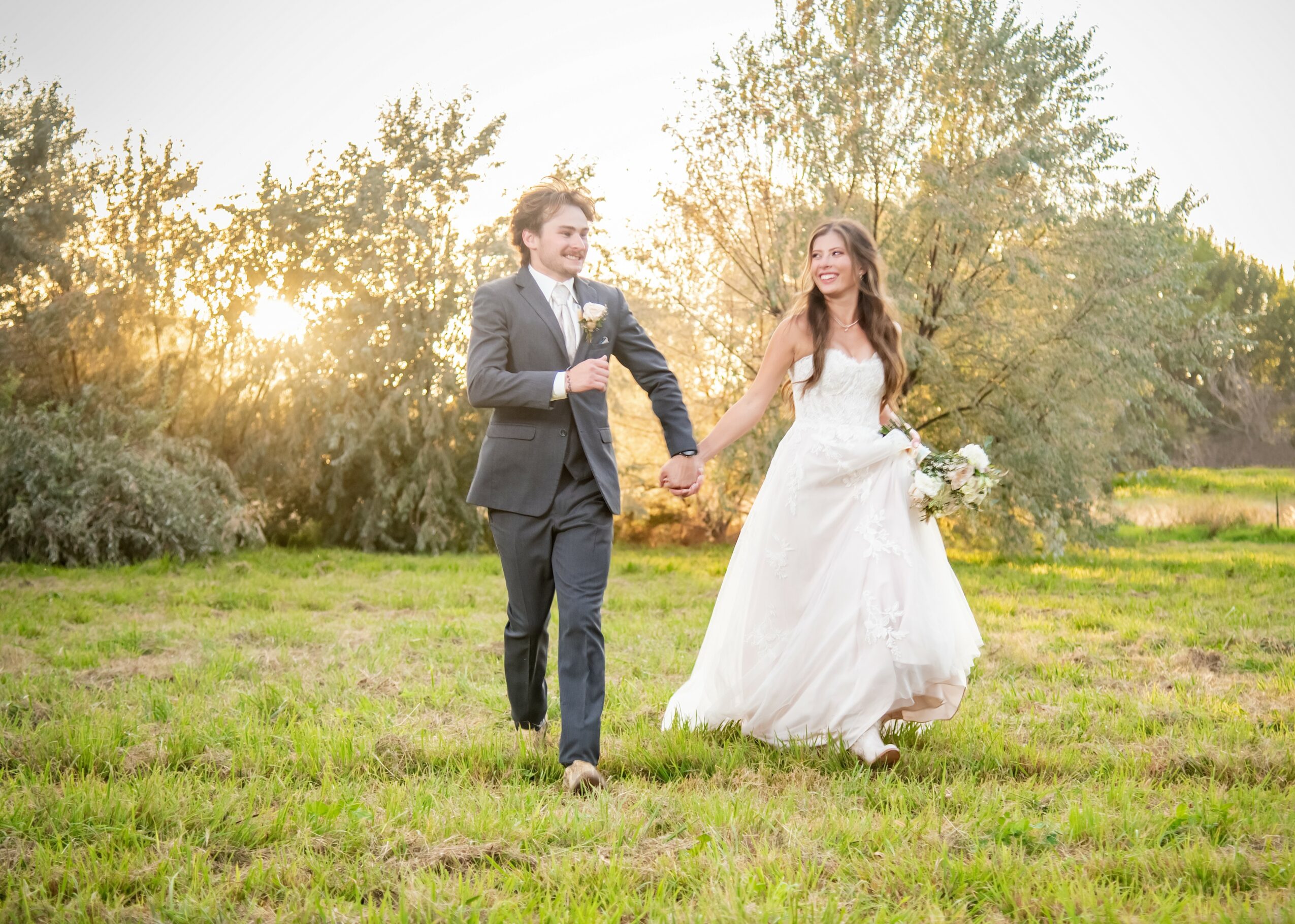 Bride and groom holding hands walking on grass in a park during sunset, trees in background.
