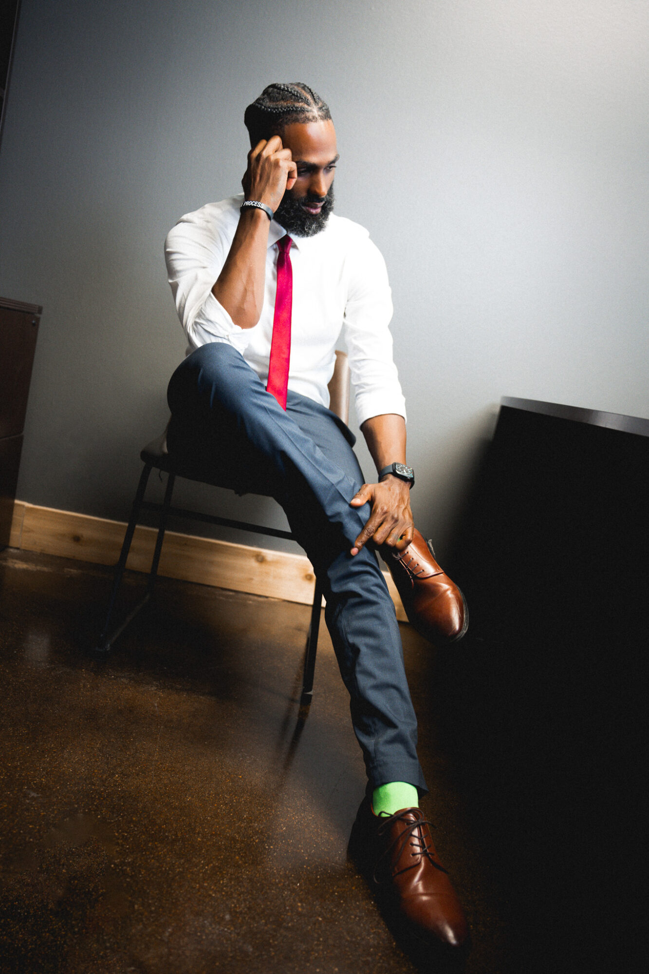 Man with beard in white shirt, red tie, dark pants, brown shoes, sitting on a chair, talking on phone.