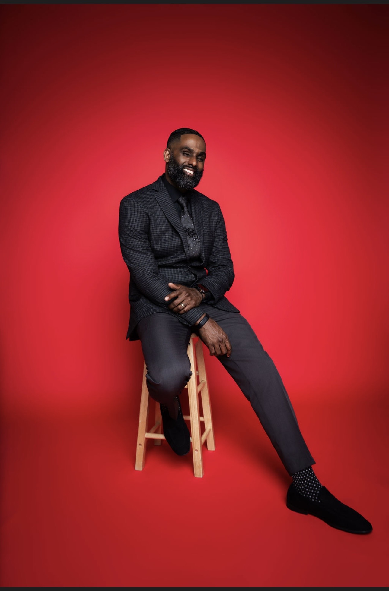 Man sitting on a stool against a red background, smiling, wearing a dark suit and black shoes.