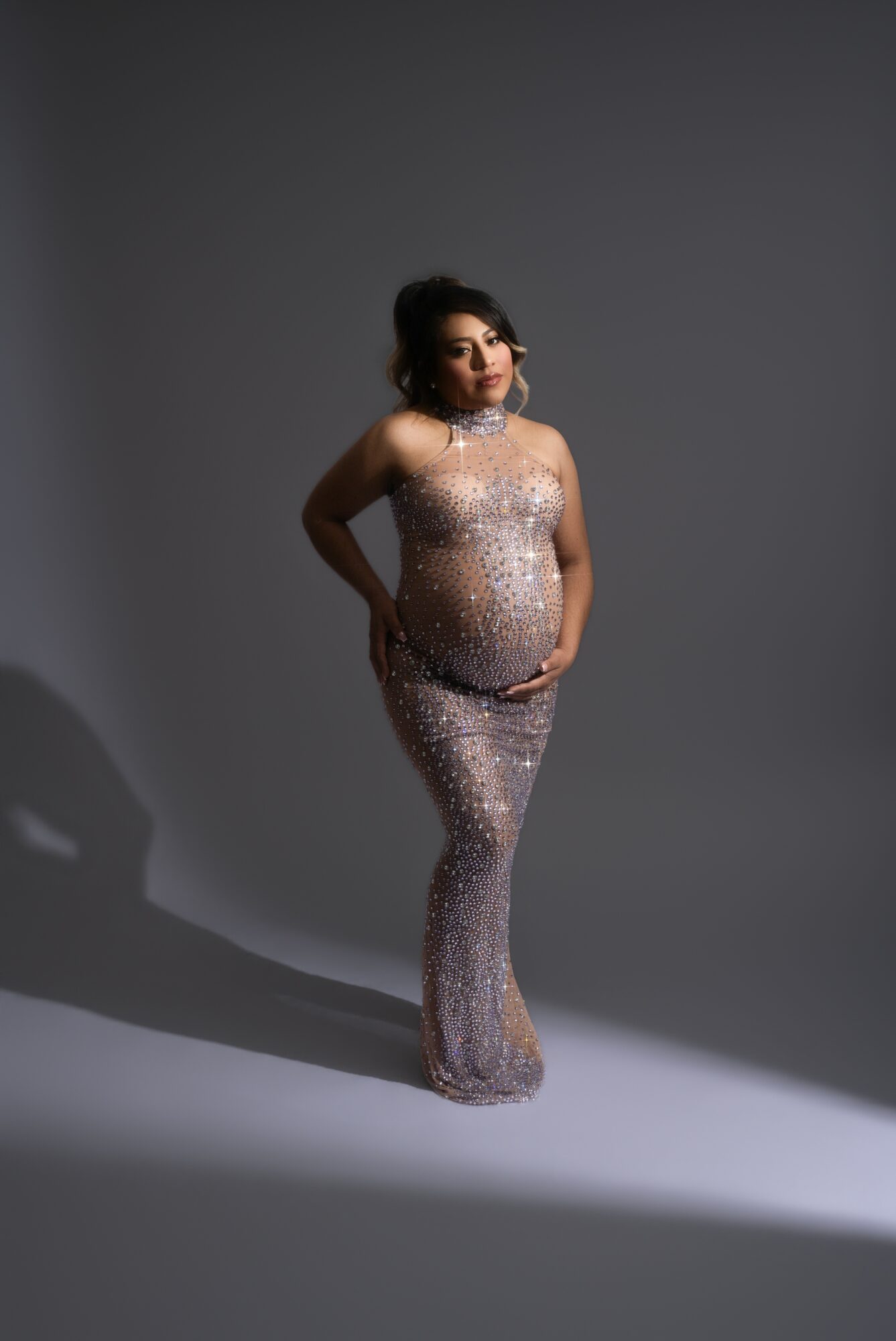 Woman in a sparkling, form-fitting gown standing against a plain gray background, with her hands on her belly.