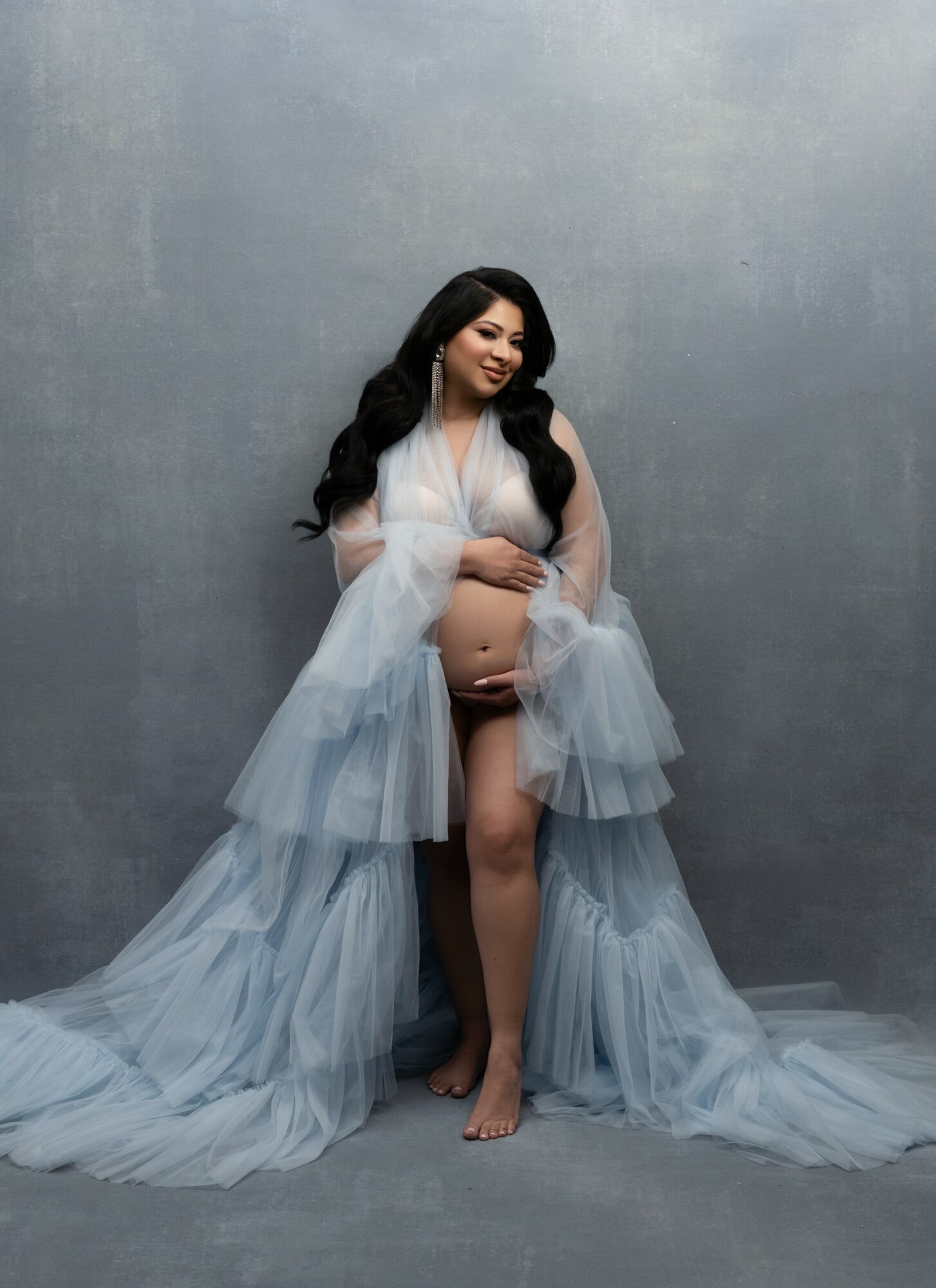 Pregnant woman with long dark hair in flowing light blue gown, standing barefoot against gray background.