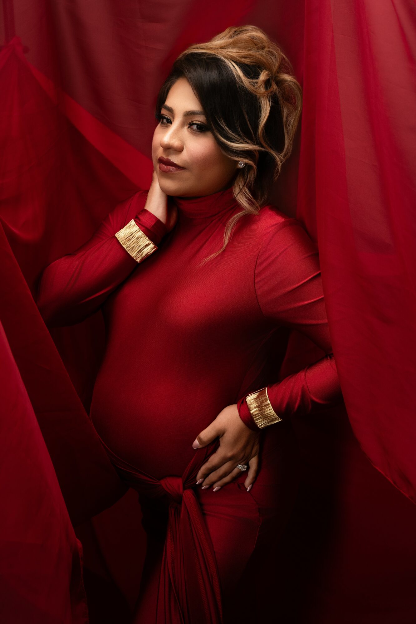 Pregnant woman with styled hair and makeup, wearing a red dress, standing among red curtains.