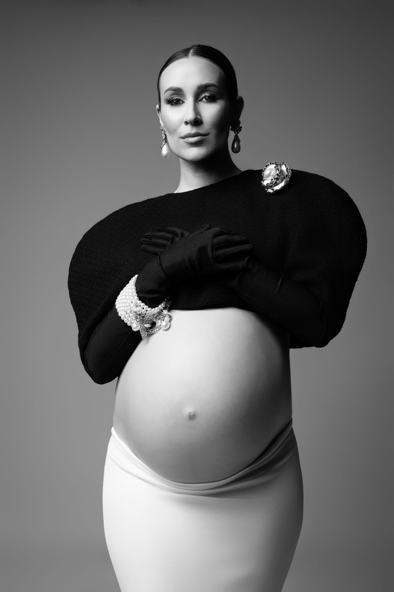 Pregnant woman in black sweater and white skirt, wearing gloves and jewelry, posing with hands on her belly.
