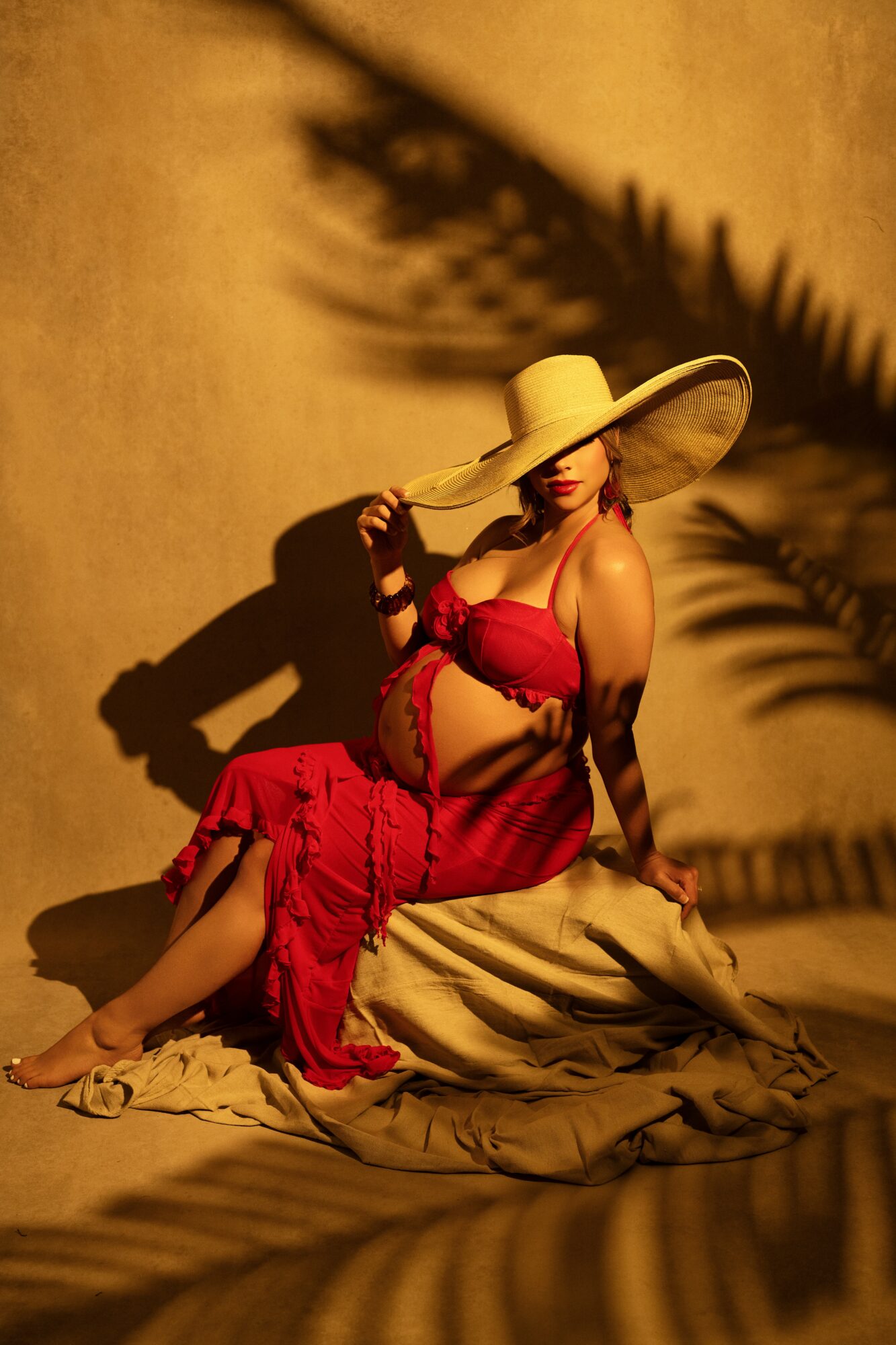 Woman in red dress and large hat sitting on fabric, shadow of palm leaves on wall behind her.