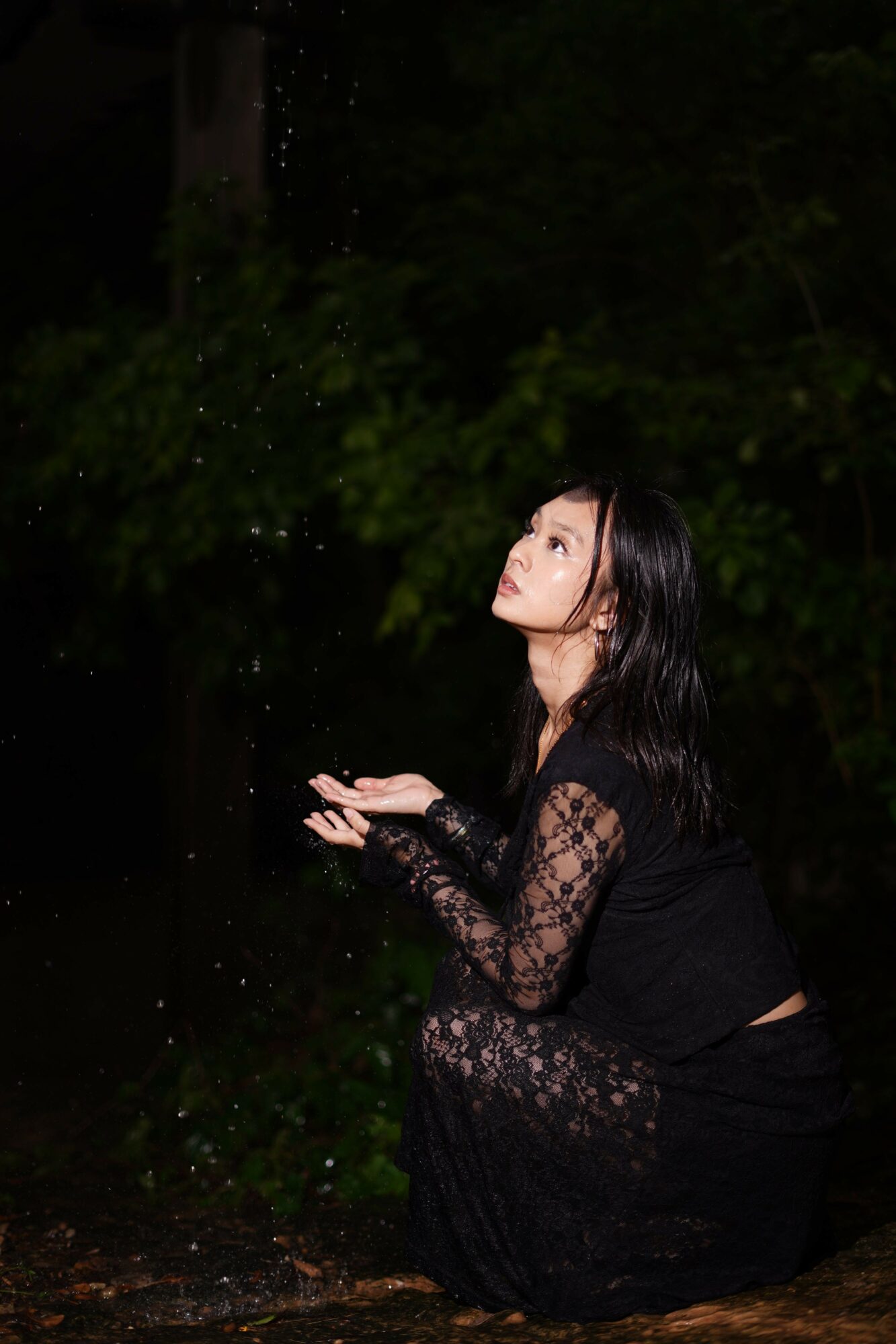 Woman kneeling outdoors at night, looking upward, with dark trees in background, wearing black lace clothing, holding small particles.