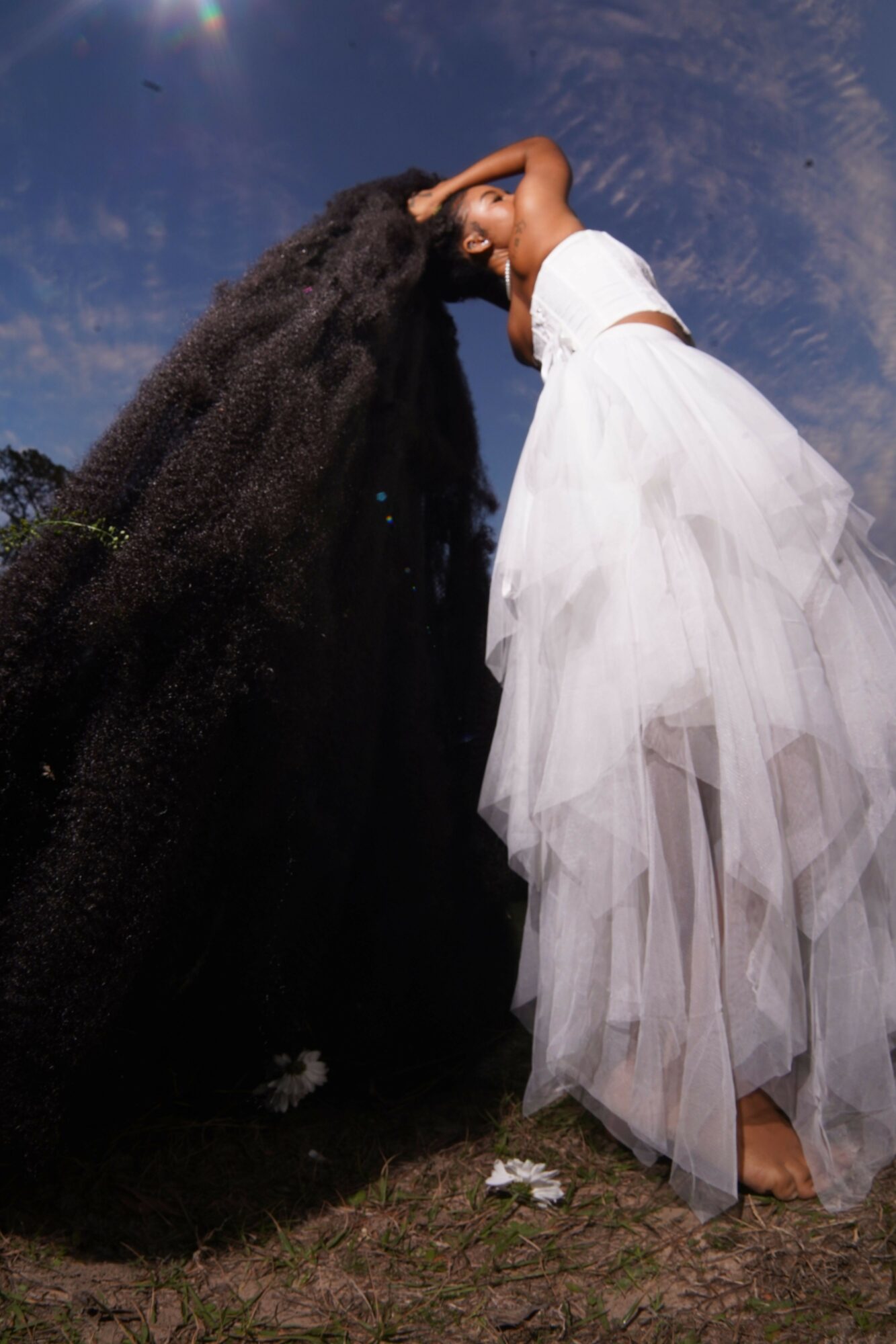 Two women in contrasting dresses stand outdoors, touching foreheads under a blue sky with clouds.