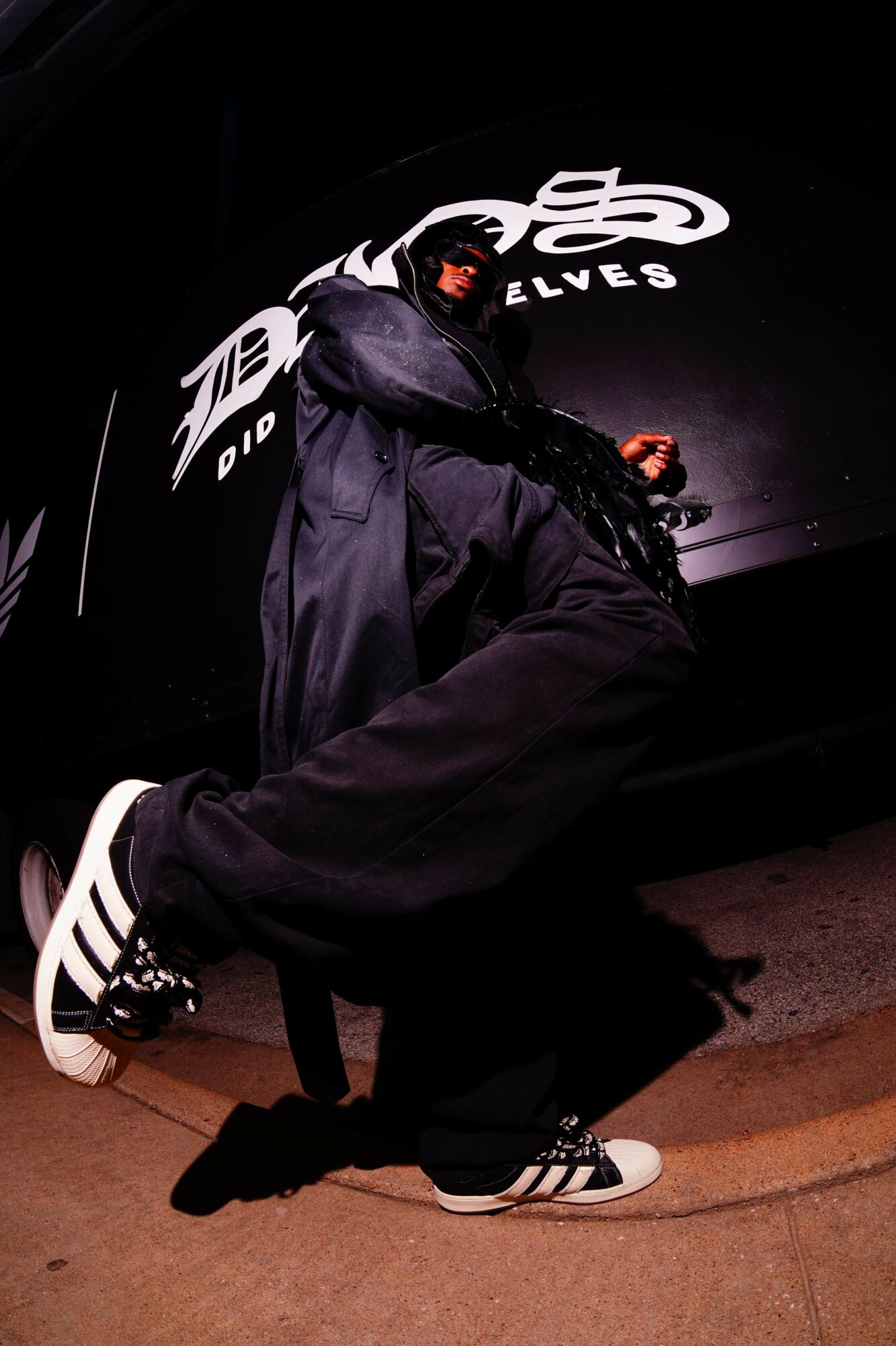 Person sitting on the ground wearing black clothing and white sneakers, with a dark background and logo behind.