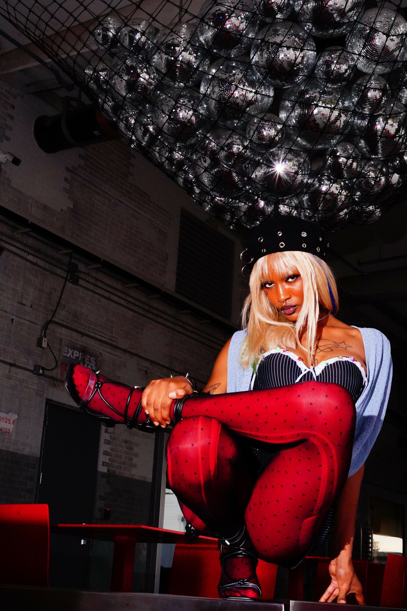 Person with blonde hair and makeup sitting on a chair, wearing red stockings and black shoes, under a chandelier.