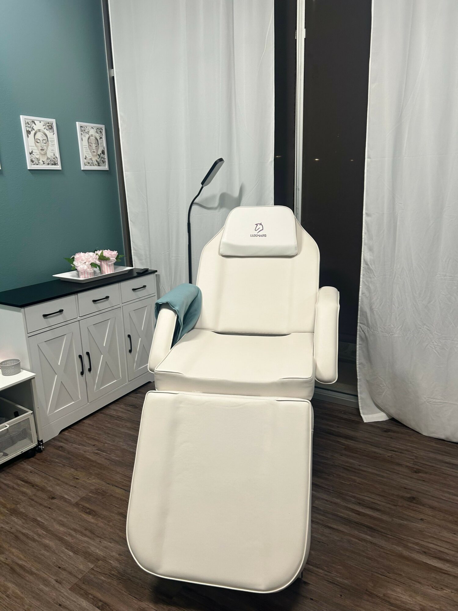 Medical or spa chair in a room with white curtains and a cabinet, with a lamp and decorative items nearby.