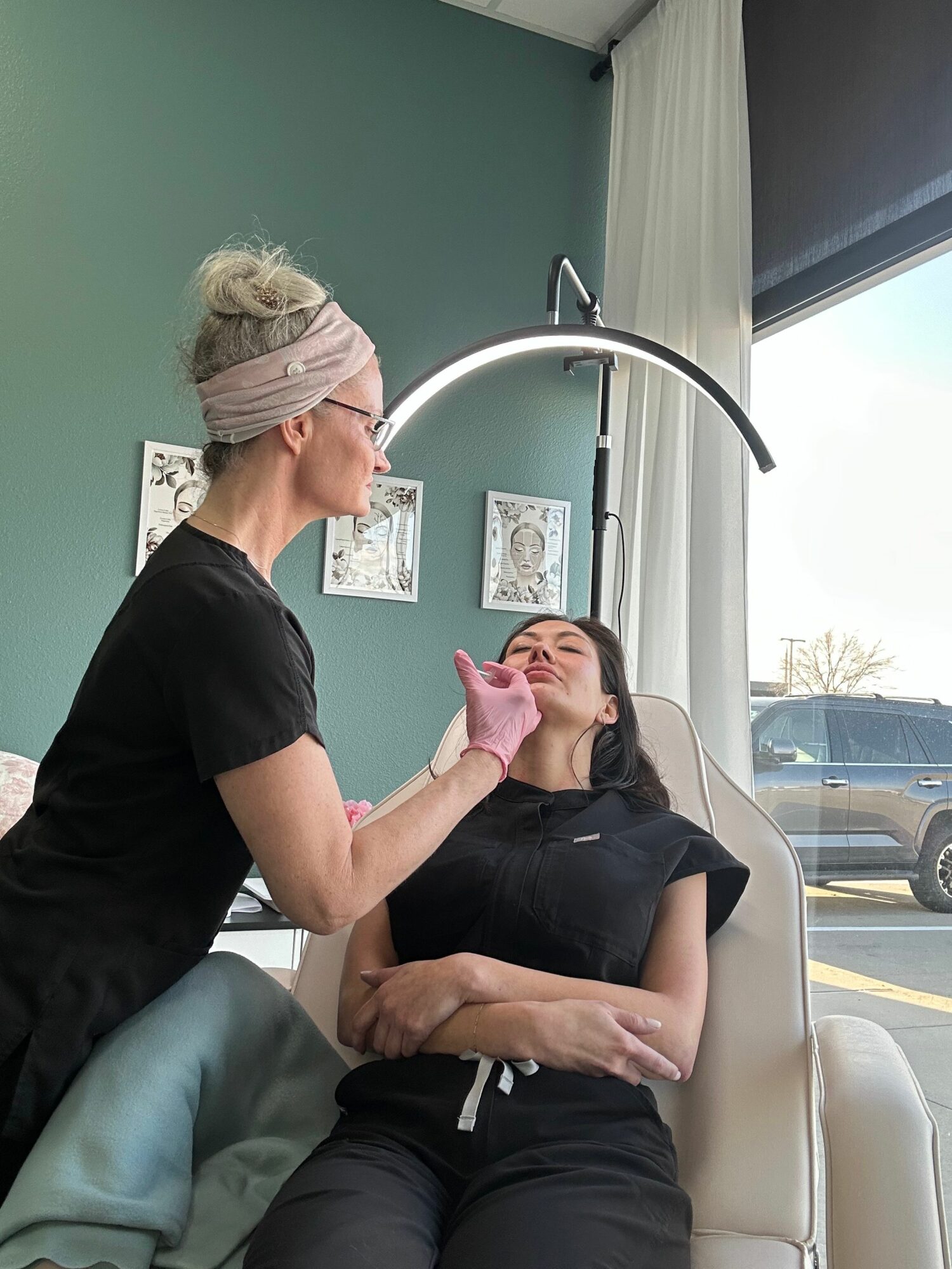 Medical professional examining a woman's face in a clinic room with teal walls and framed artwork.