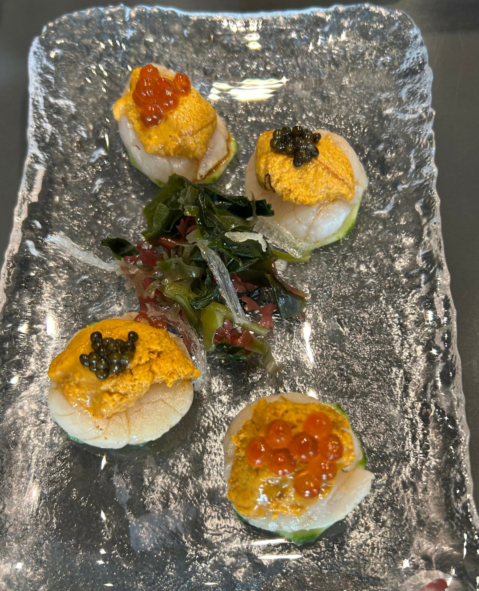 Four pieces of sushi with yellow topping and black or orange garnish, arranged on a textured glass plate with seaweed in the center.
