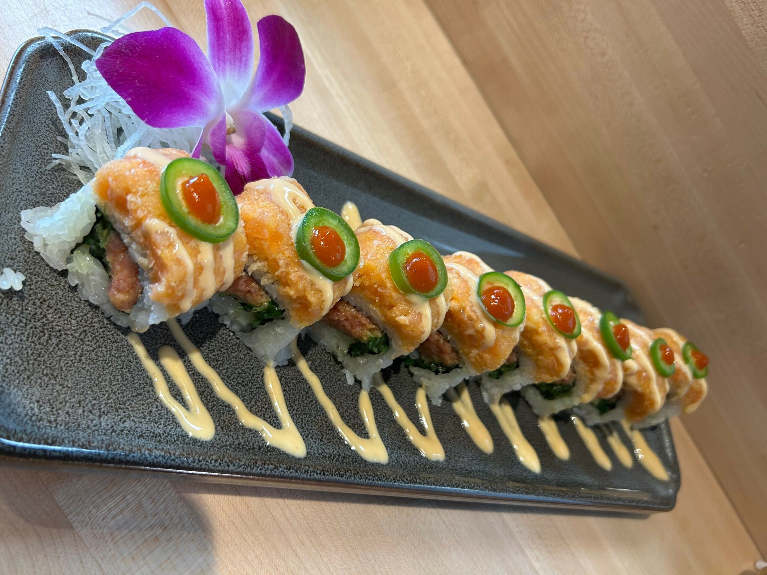 Sushi rolls with orange sauce, green garnish, and purple flower on black rectangular plate, drizzled with yellow sauce.