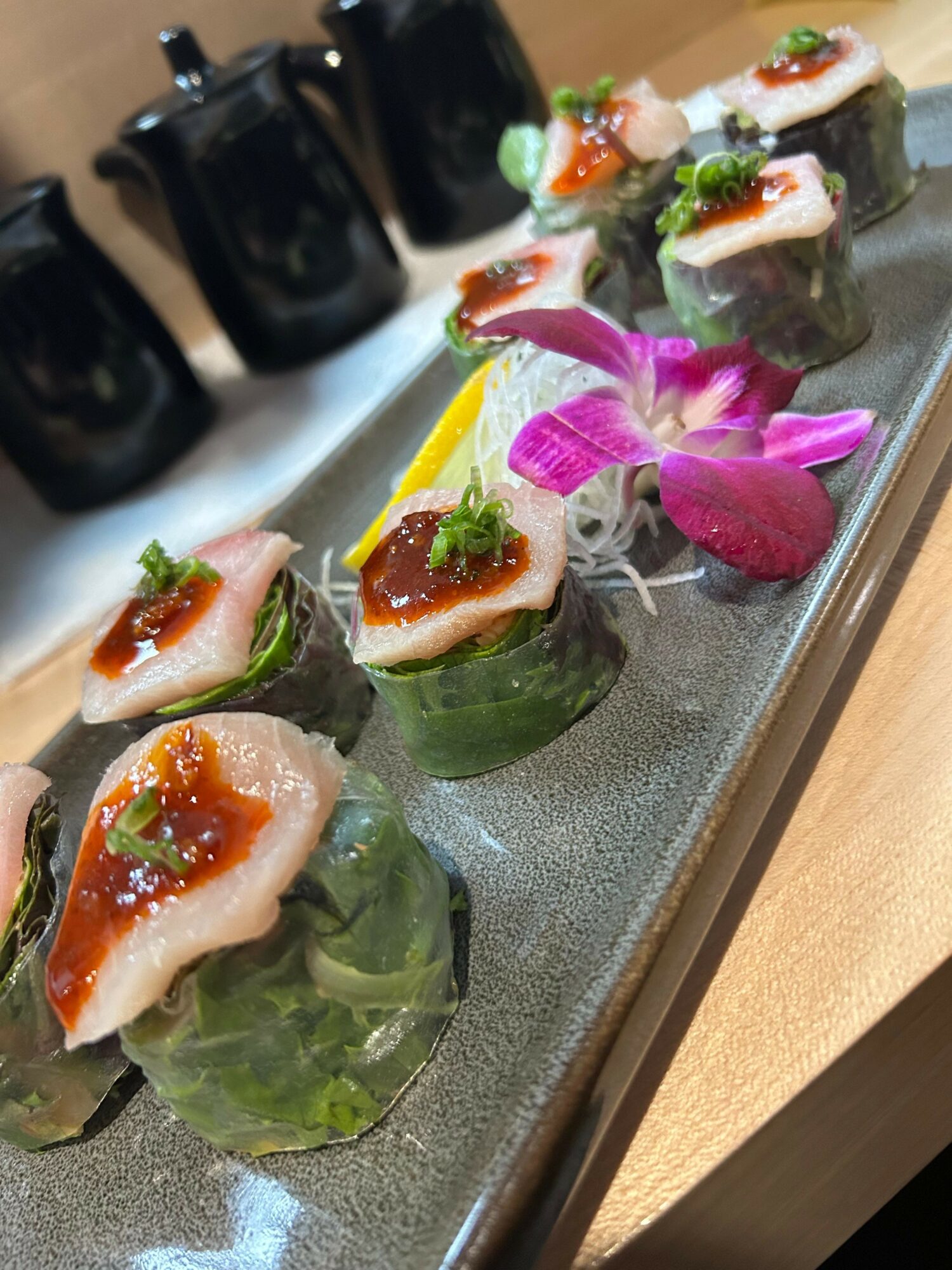 Assorted sushi rolls with fish, garnished with green herbs and pink flowers on a black rectangular plate.
