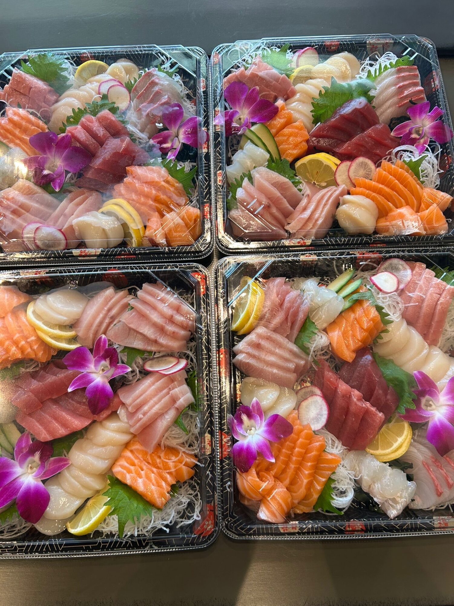 Four containers of assorted sushi with colorful fish, shrimp, and garnishes, decorated with purple flowers.