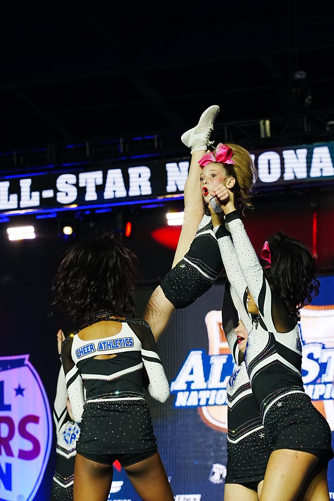 Two young girls in cheerleading outfits perform a routine on stage with a large All-Star banner behind them.