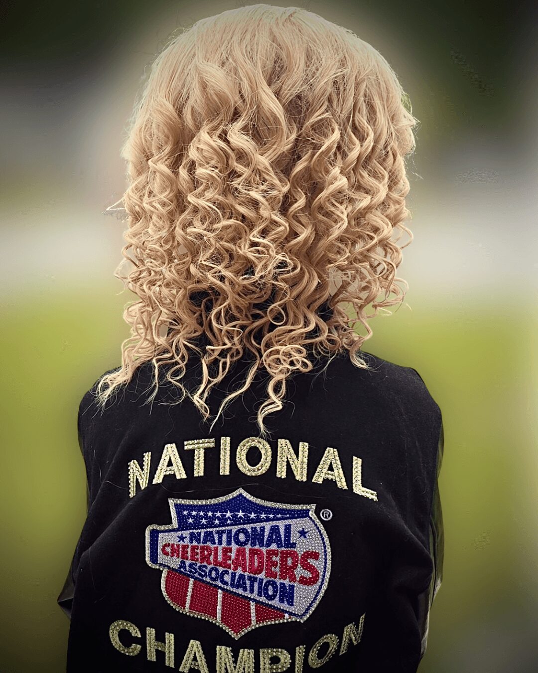 Back of a person with curly blonde hair wearing a black jacket with 'NATIONAL CHAMPION' and a cheerleading logo.