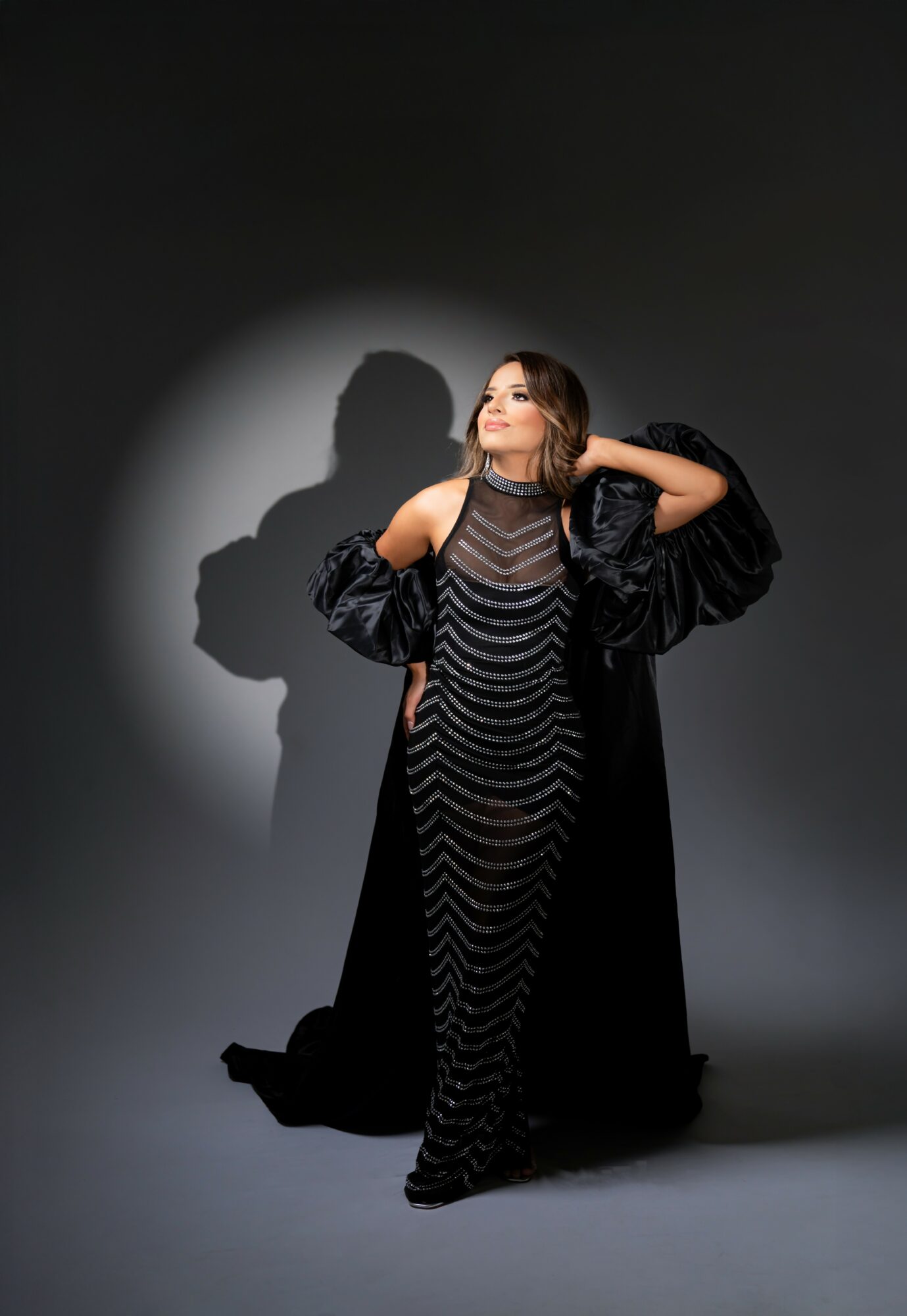 Woman in a black dress and cape posing with hands behind head, shadow cast on dark background.