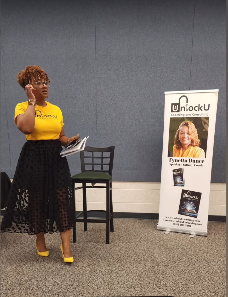 Woman with curly hair, glasses, yellow top, black skirt, yellow shoes, holding papers, standing next to a chair and a display board.