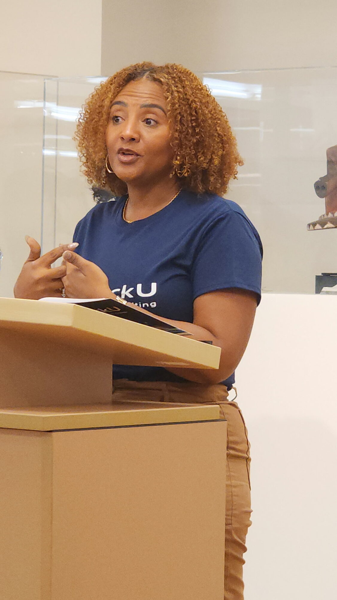 Woman with curly hair speaking at a podium in a room with a glass wall.