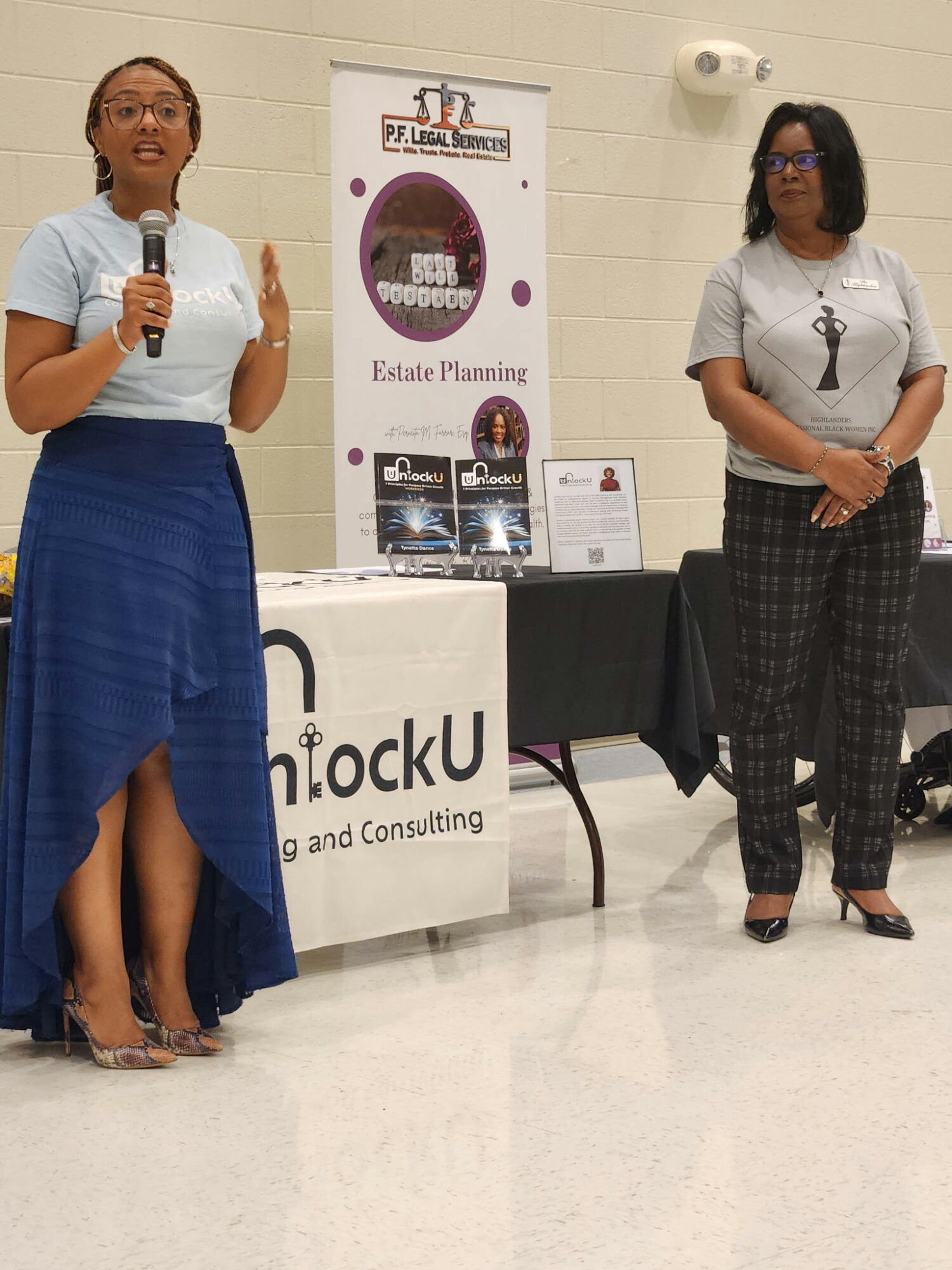 Two women standing indoors, one speaking into a microphone, the other listening, with banners and tables behind them.