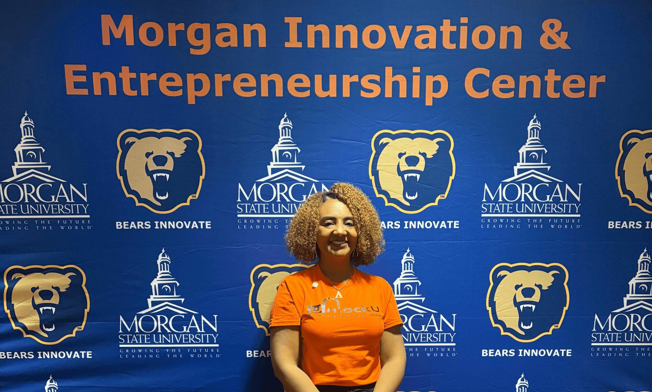 Woman with curly hair smiling in front of a blue backdrop with text and logos for Morgan Innovation & Entrepreneurship Center.