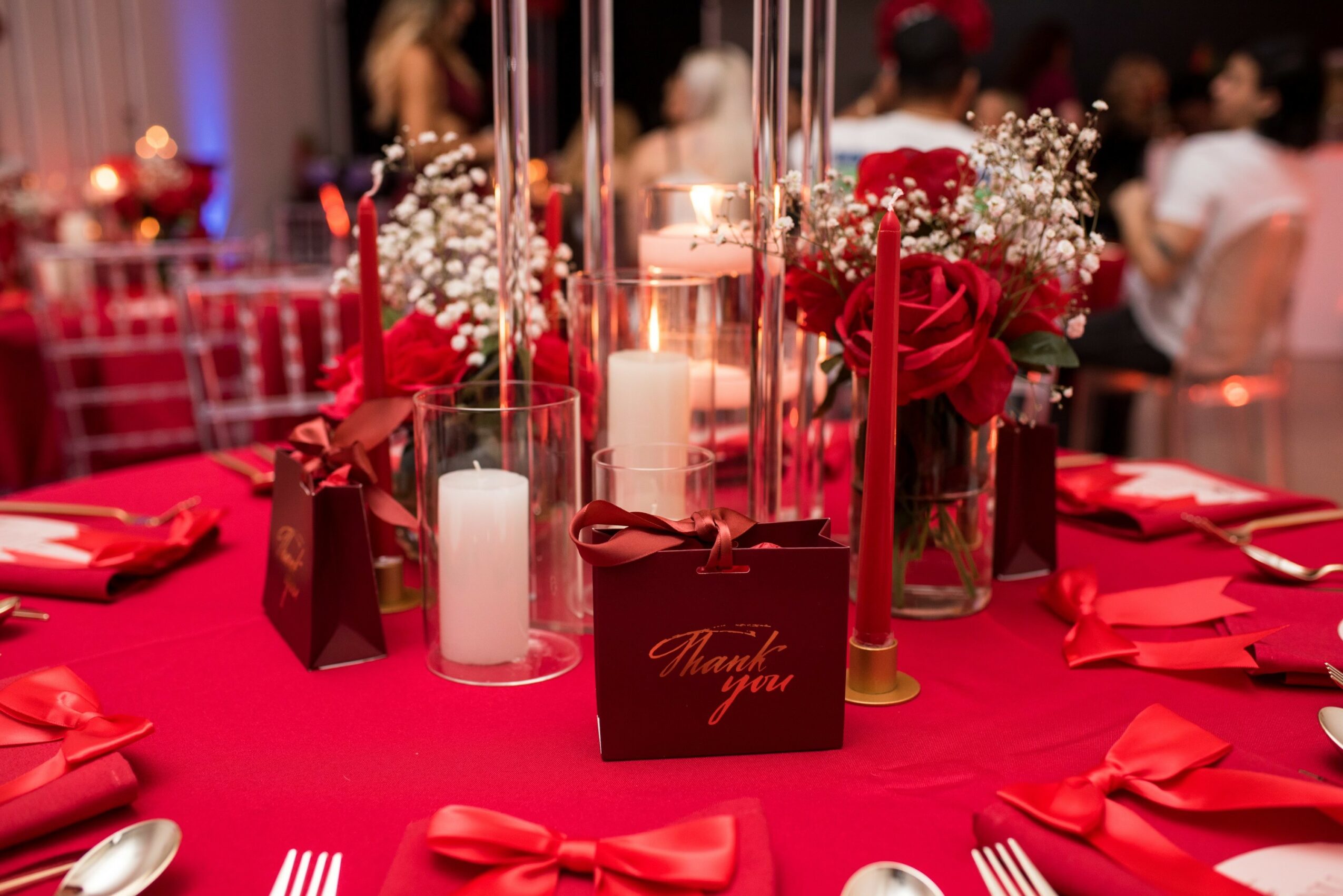 Table decorated with red candles, flowers, and gift bags at a festive event, with people in the background.