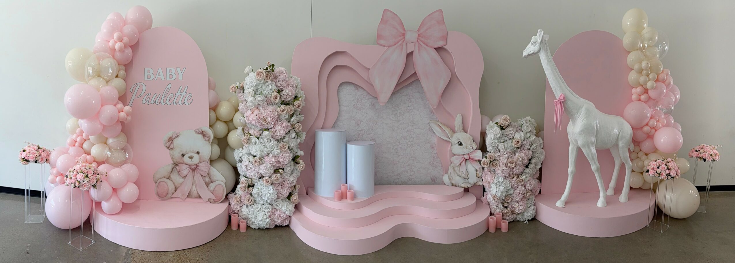 Decorative pink and white setup with balloons, teddy bears, a giraffe, and floral arrangements, featuring a pink heart-shaped frame.