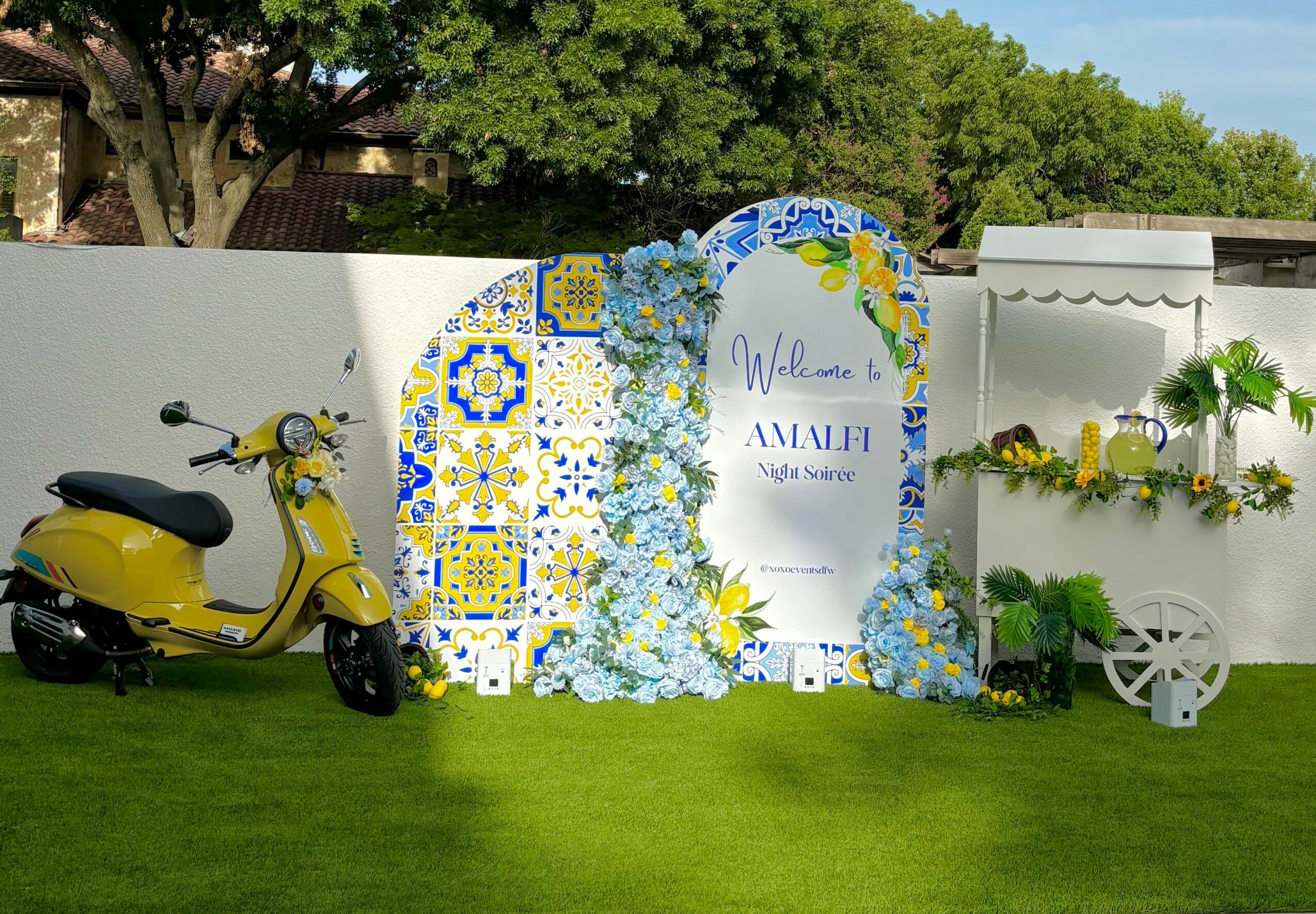 Yellow scooter parked on grass next to decorative arches with blue and yellow tiles and flowers, and a white cart with plants.