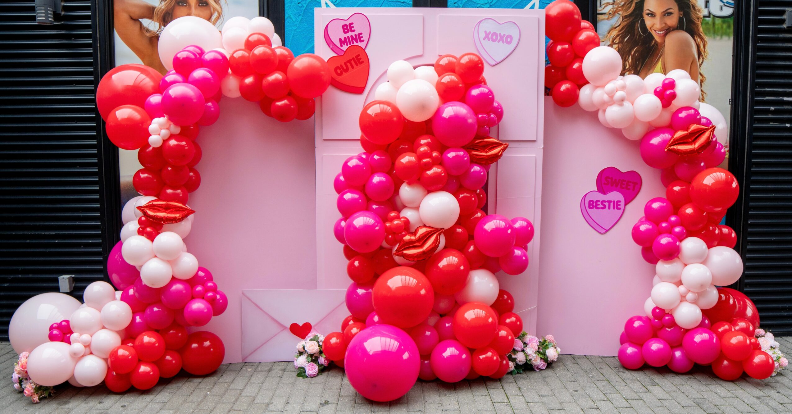 Colorful balloon arrangement with pink, red, and white balloons, and heart-shaped signs, in front of a white backdrop with a woman in the background.