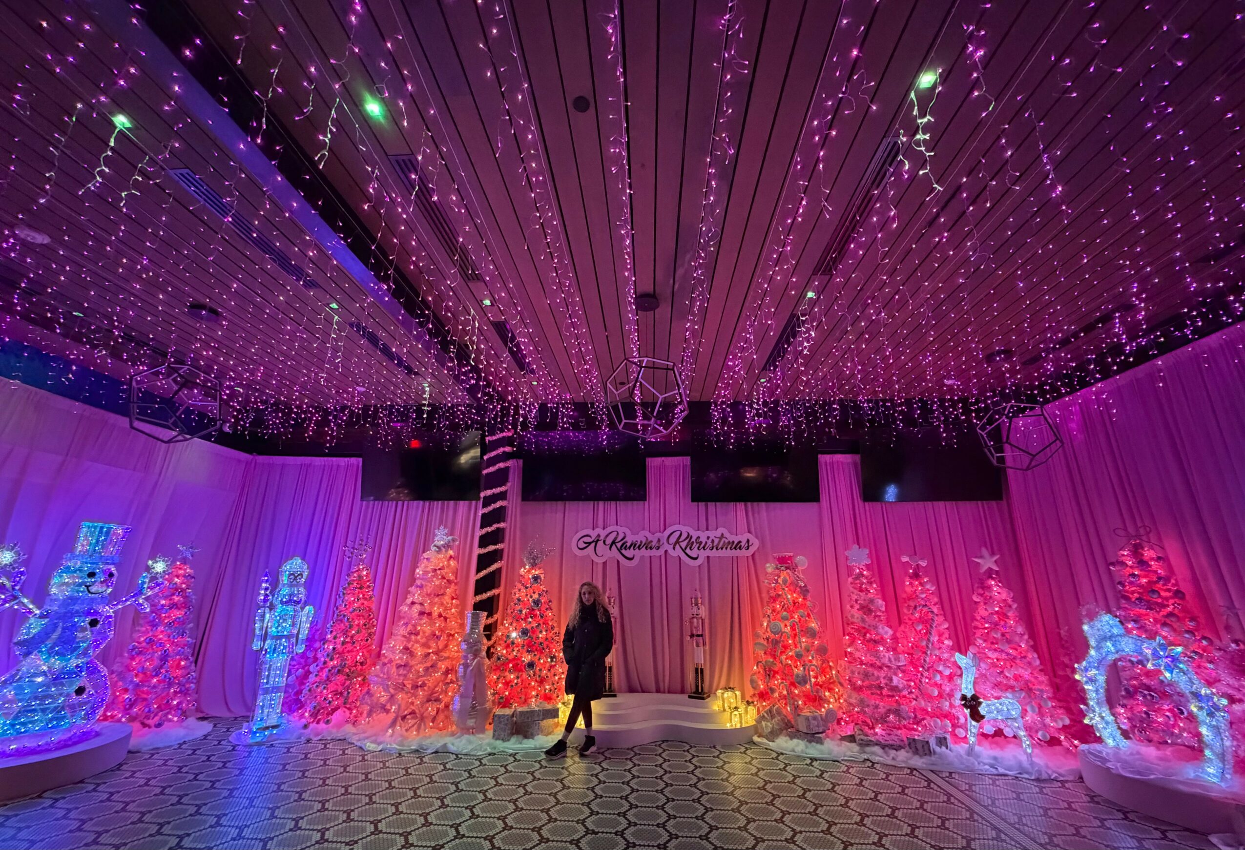 Indoor event space decorated with colorful illuminated Christmas trees and reindeer figures, with a person walking in the center.
