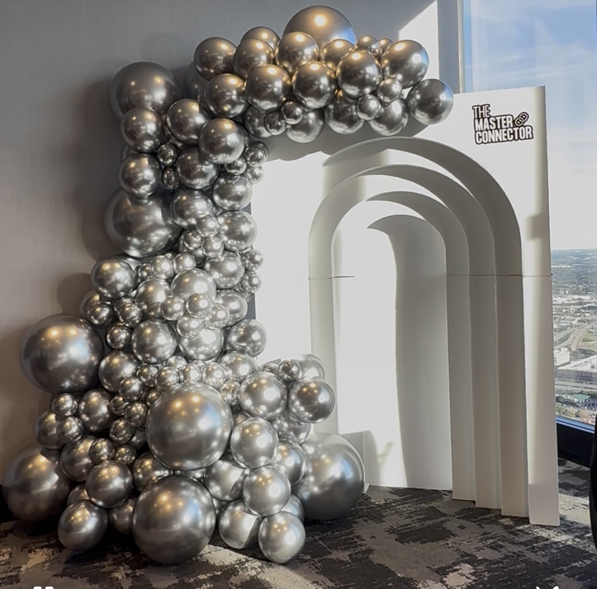 Archway decorated with silver balloons in front of a white backdrop with a city view outside window.