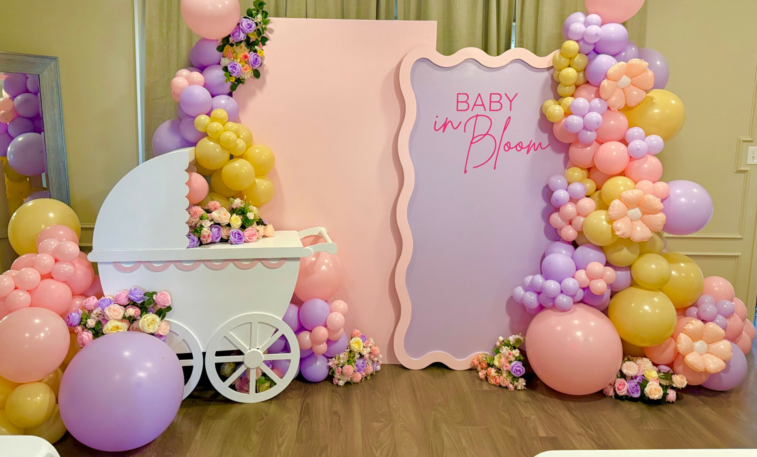 Decorative backdrop with pastel balloons, a pink sign, and a white baby stroller with flowers for a baby shower.