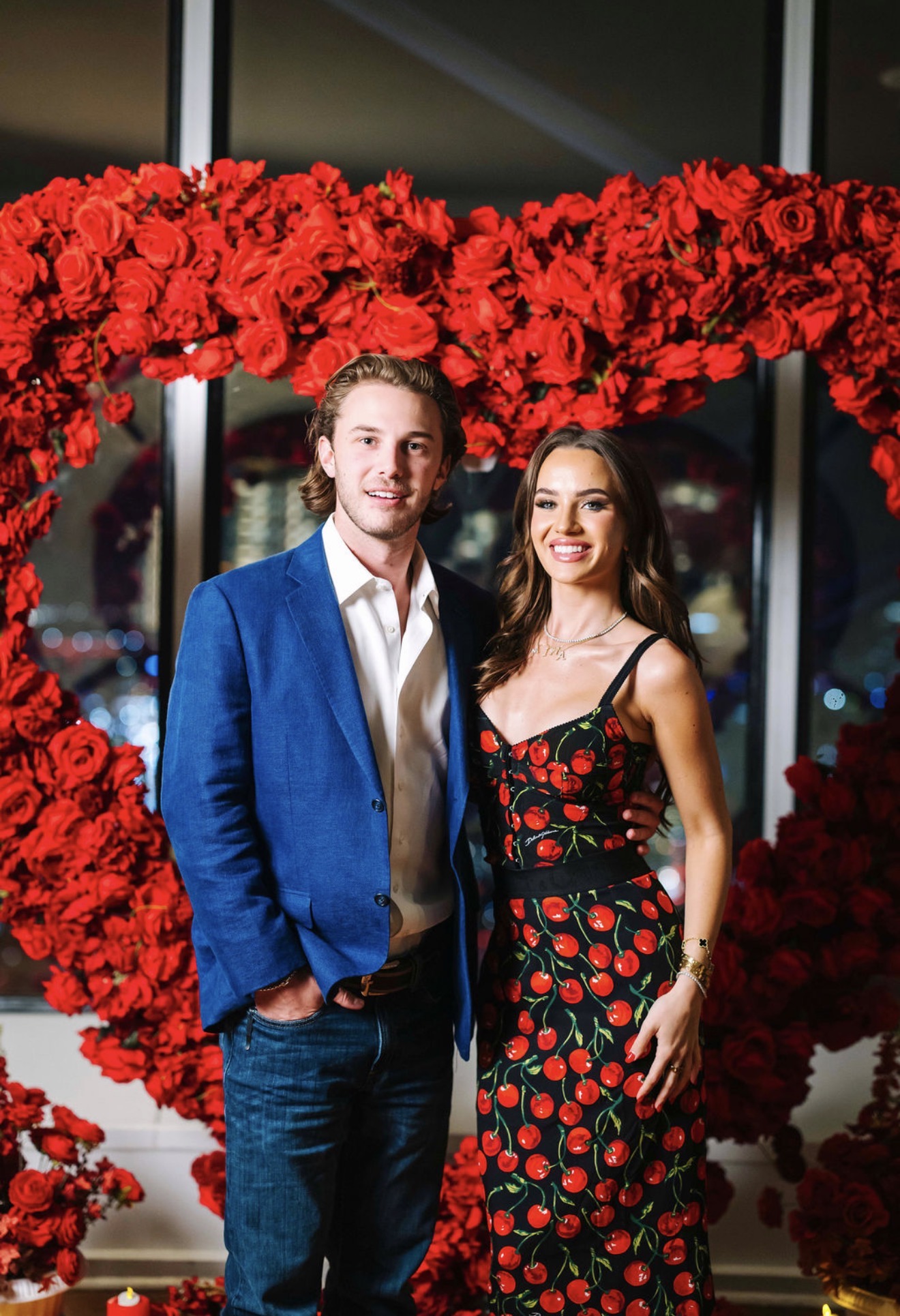Man and woman stand together in front of a heart-shaped floral display of red flowers, smiling at the camera.