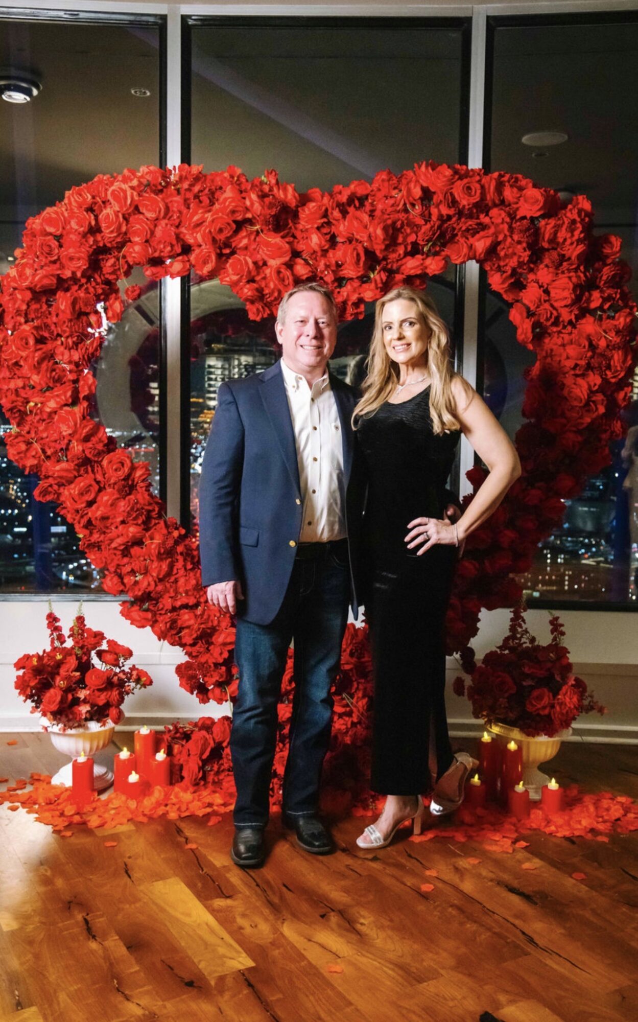 Man and woman stand inside a heart-shaped flower arrangement with city skyline in background.