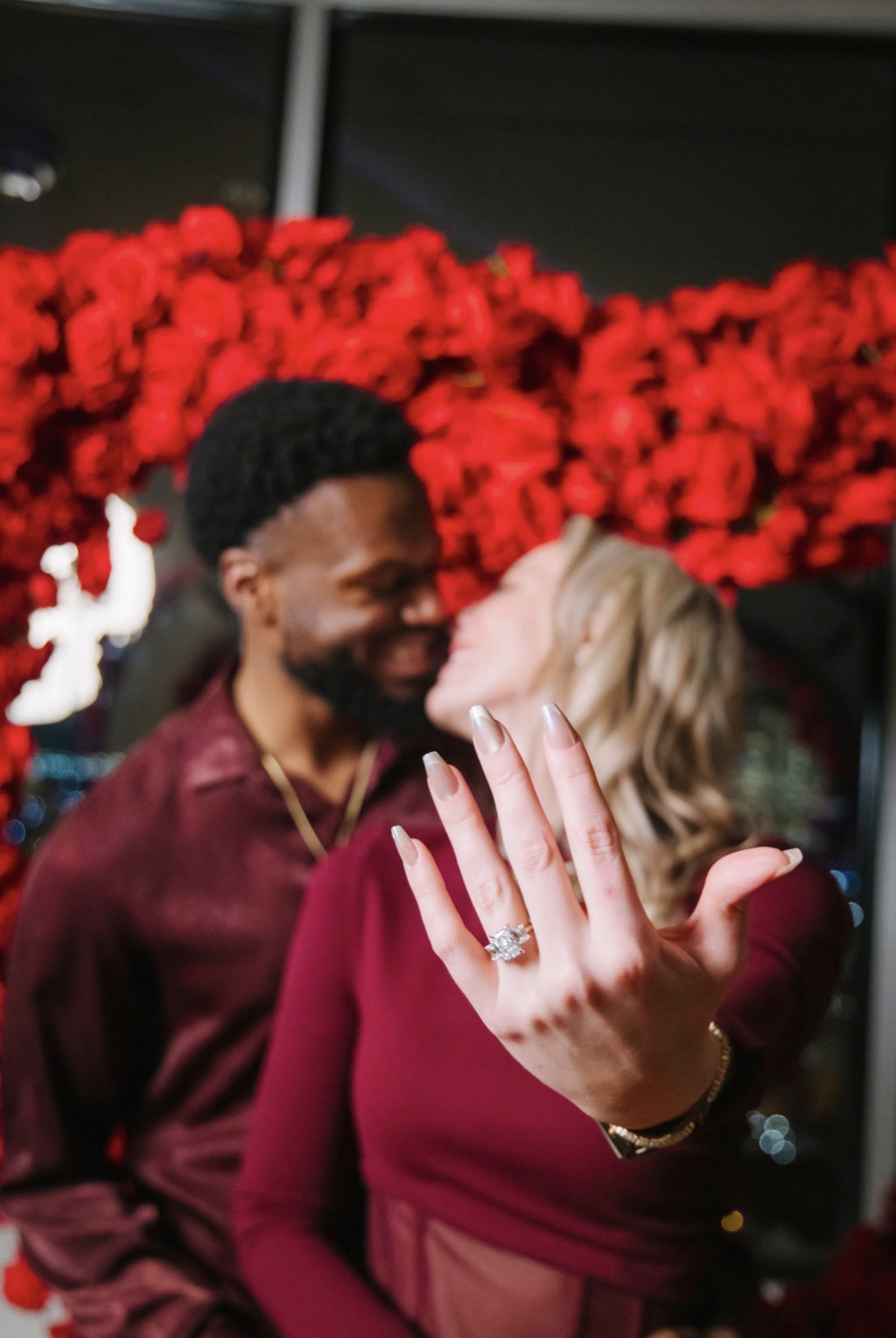 A woman shows her engagement ring while a man looks at her smiling, with red flowers in the background.