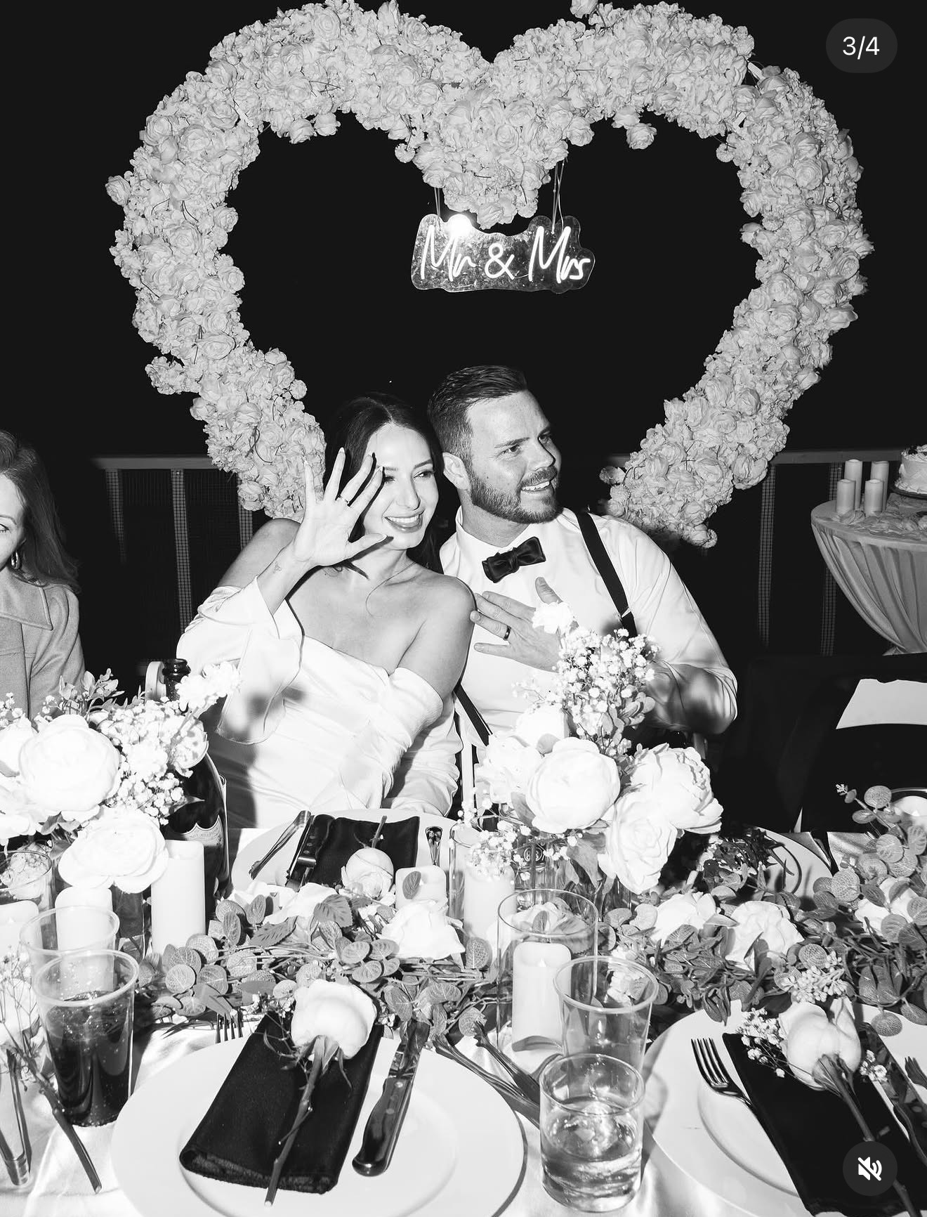 Bride and groom sitting at a table with floral decorations, heart-shaped flower arrangement, and a sign reading 'Mr & Mrs'.