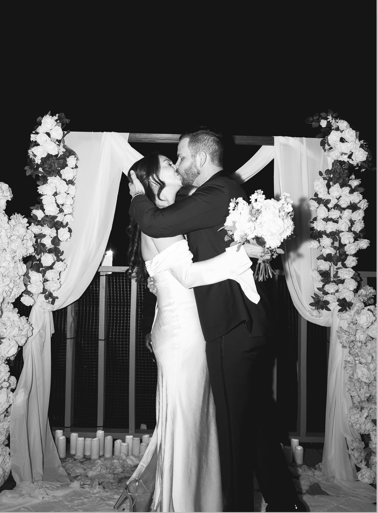 Couple kissing at wedding, holding bouquet, surrounded by floral decorations and curtains.