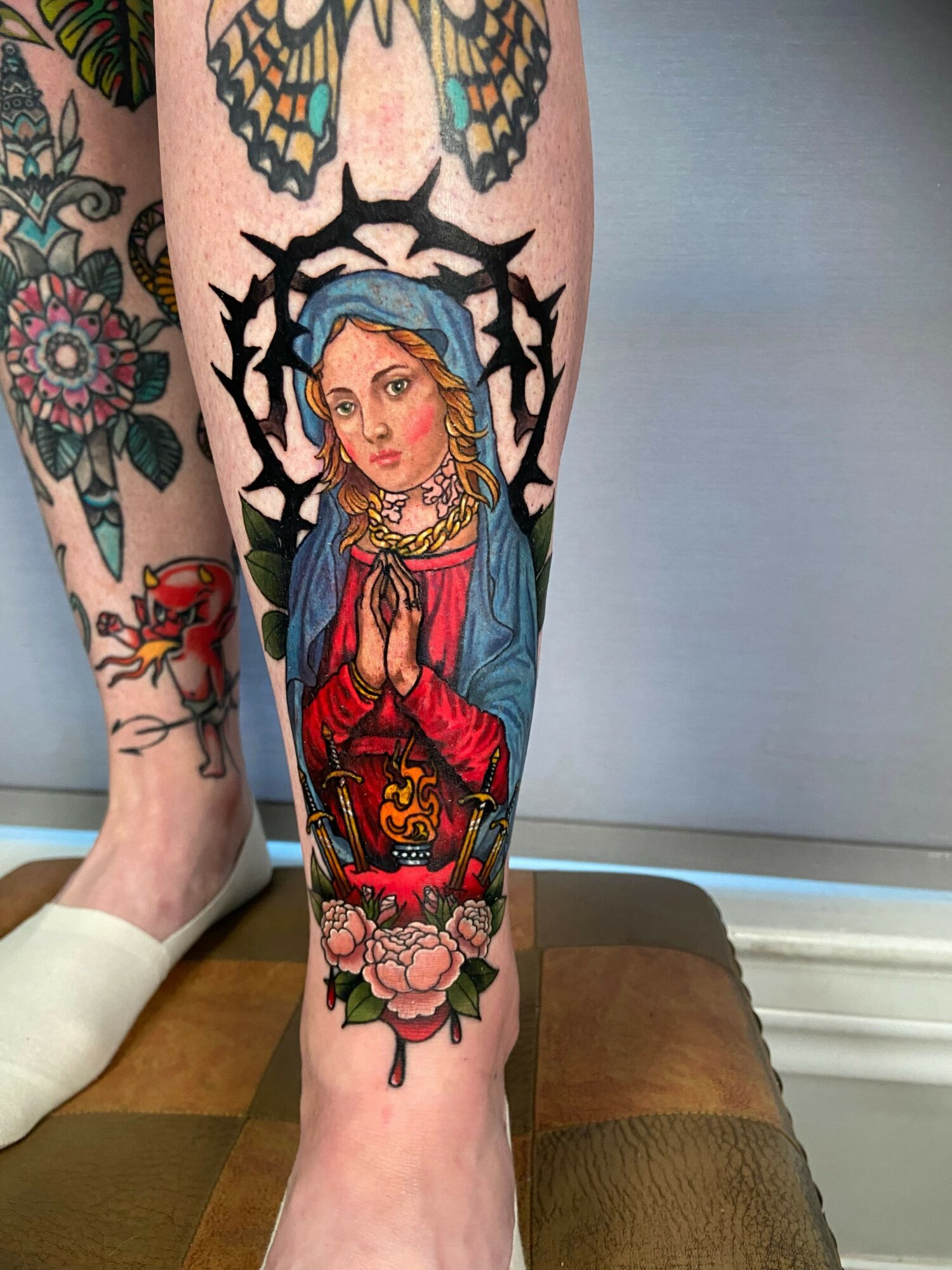 Tattoo of a woman with a blue headscarf, praying hands, surrounded by flowers and a crown, on a person's leg.