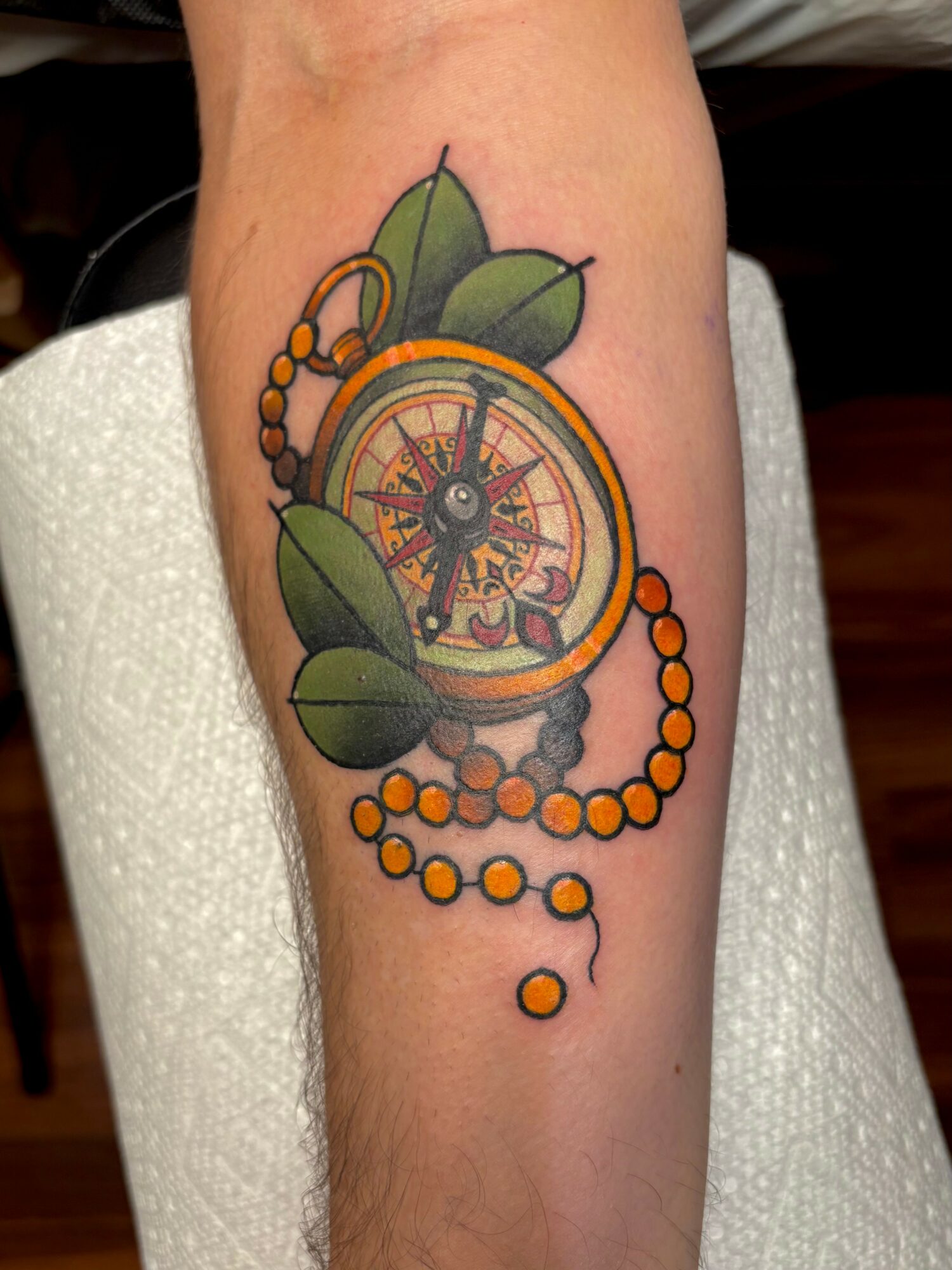 Tattoo of a compass with leaves and a beaded chain on a person's arm.
