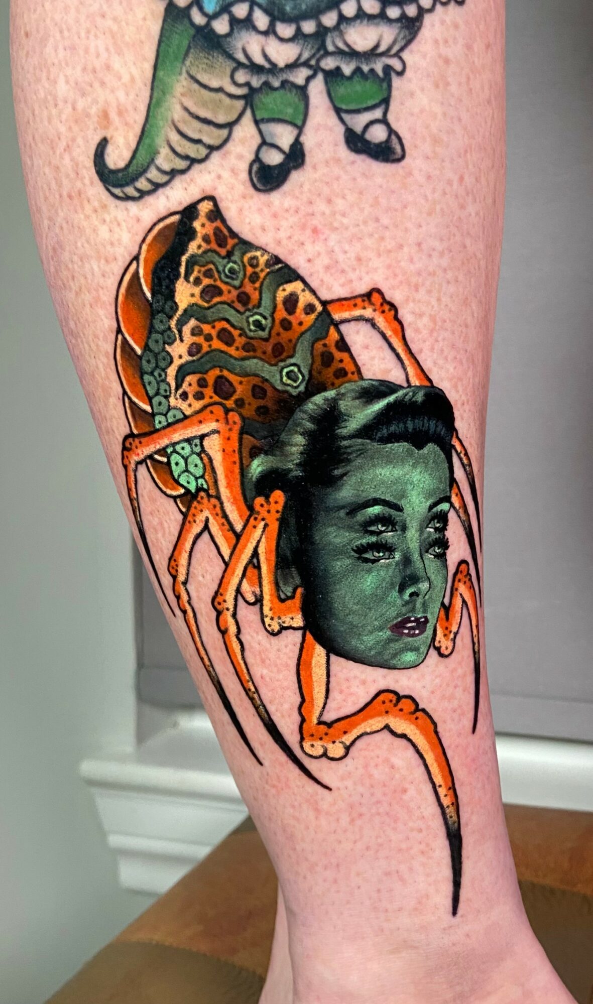 Tattoo of a spider with a human face, detailed orange and black body, on a person's leg.