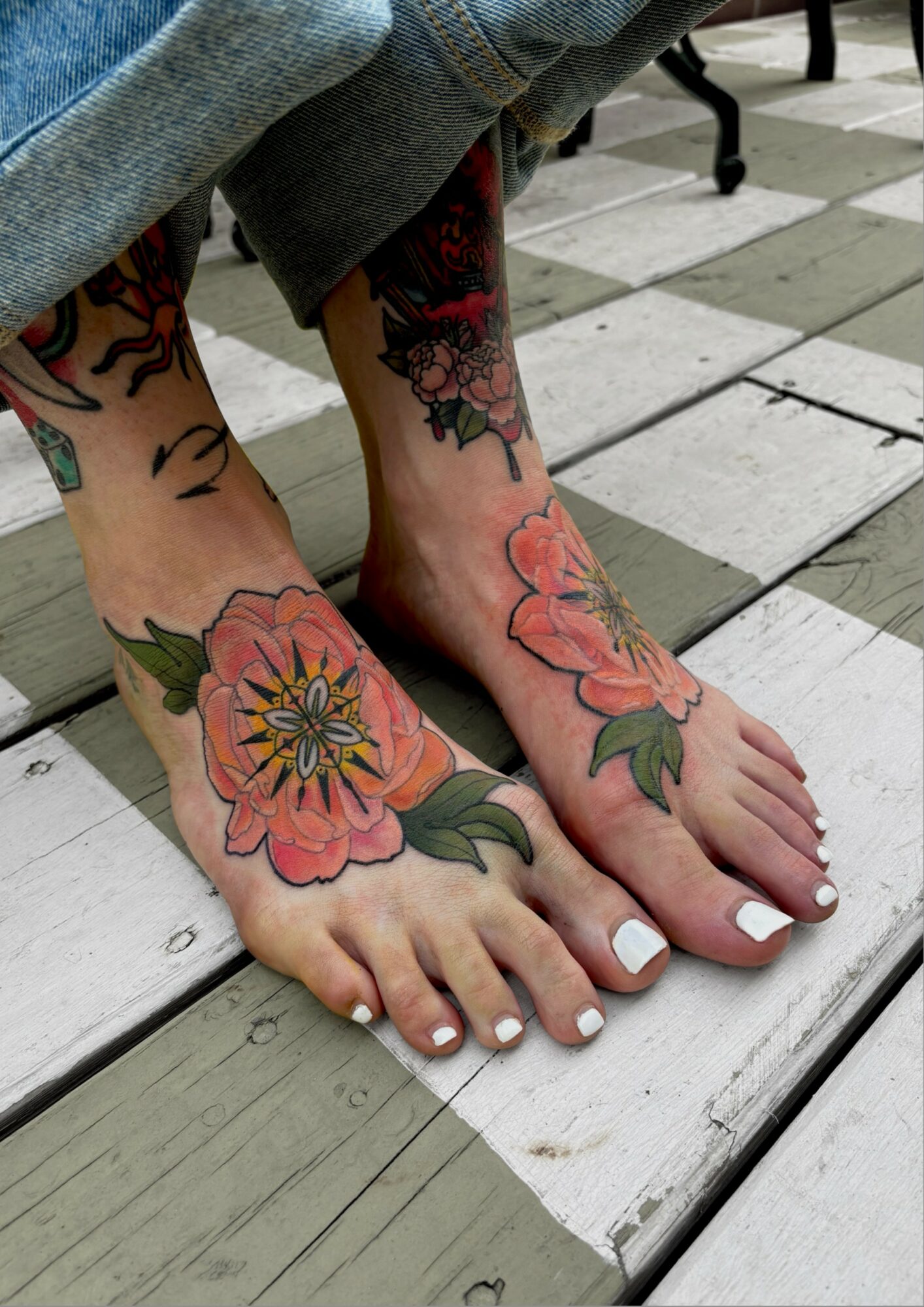 Feet with colorful floral tattoos on toes and ankles, standing on wooden and tiled surface.